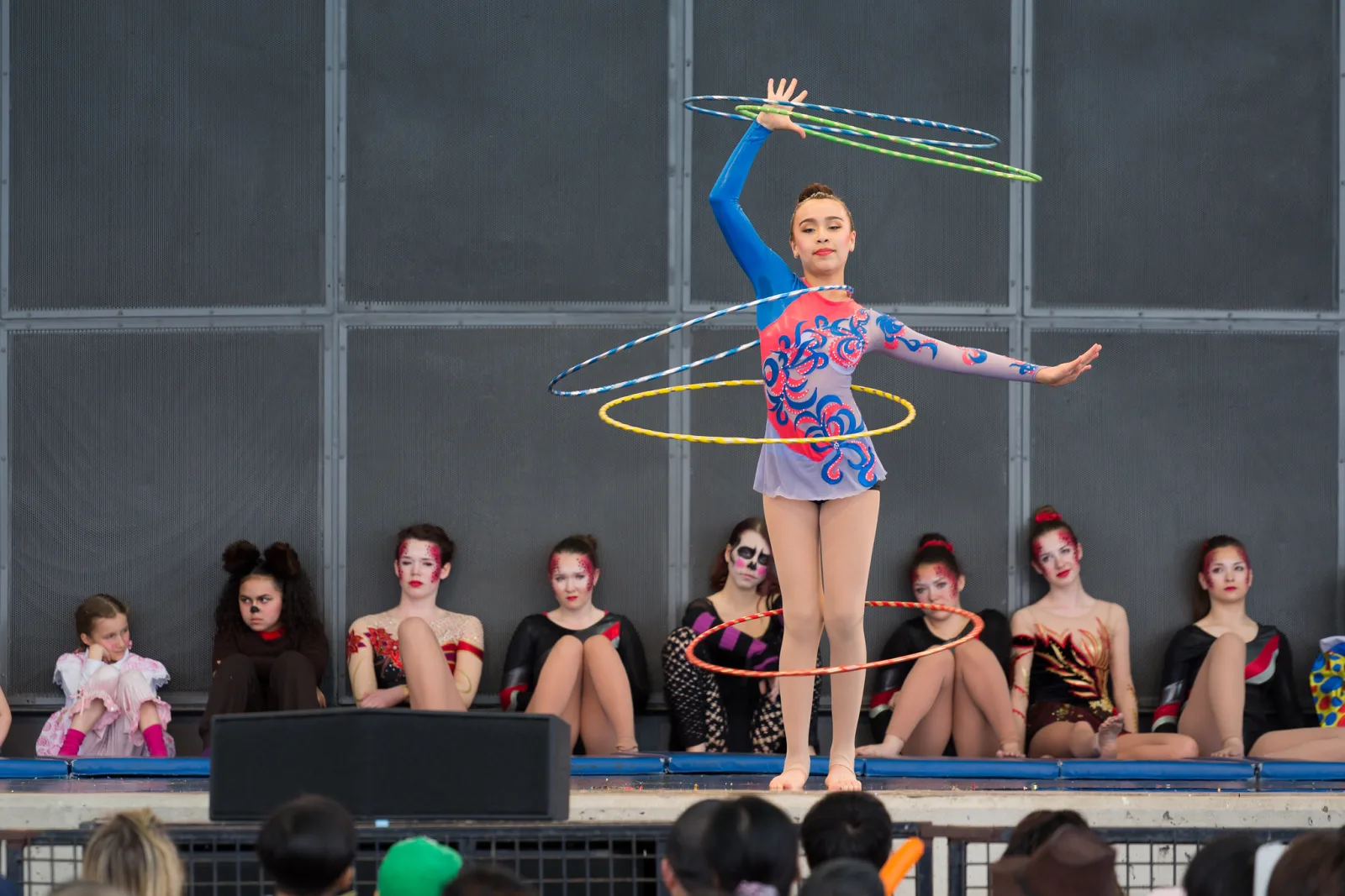 A young performer on an outdoor stage balancing on her toes with one arm raised and one extended, spinning several hoops at once, while costumed performers sit along the back of the stage