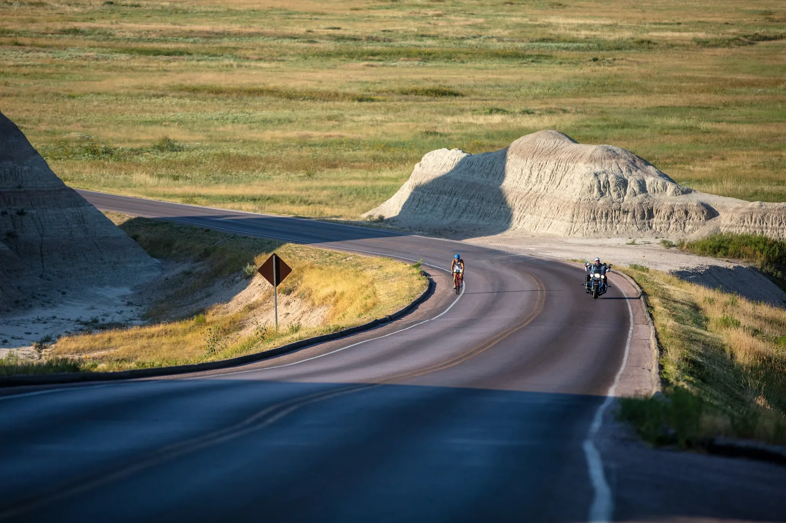 A cyclist and a motorcyclist riding toward the camera on a winding road through grassland and pale eroded hills, with a long empty curve of road in the foreground
