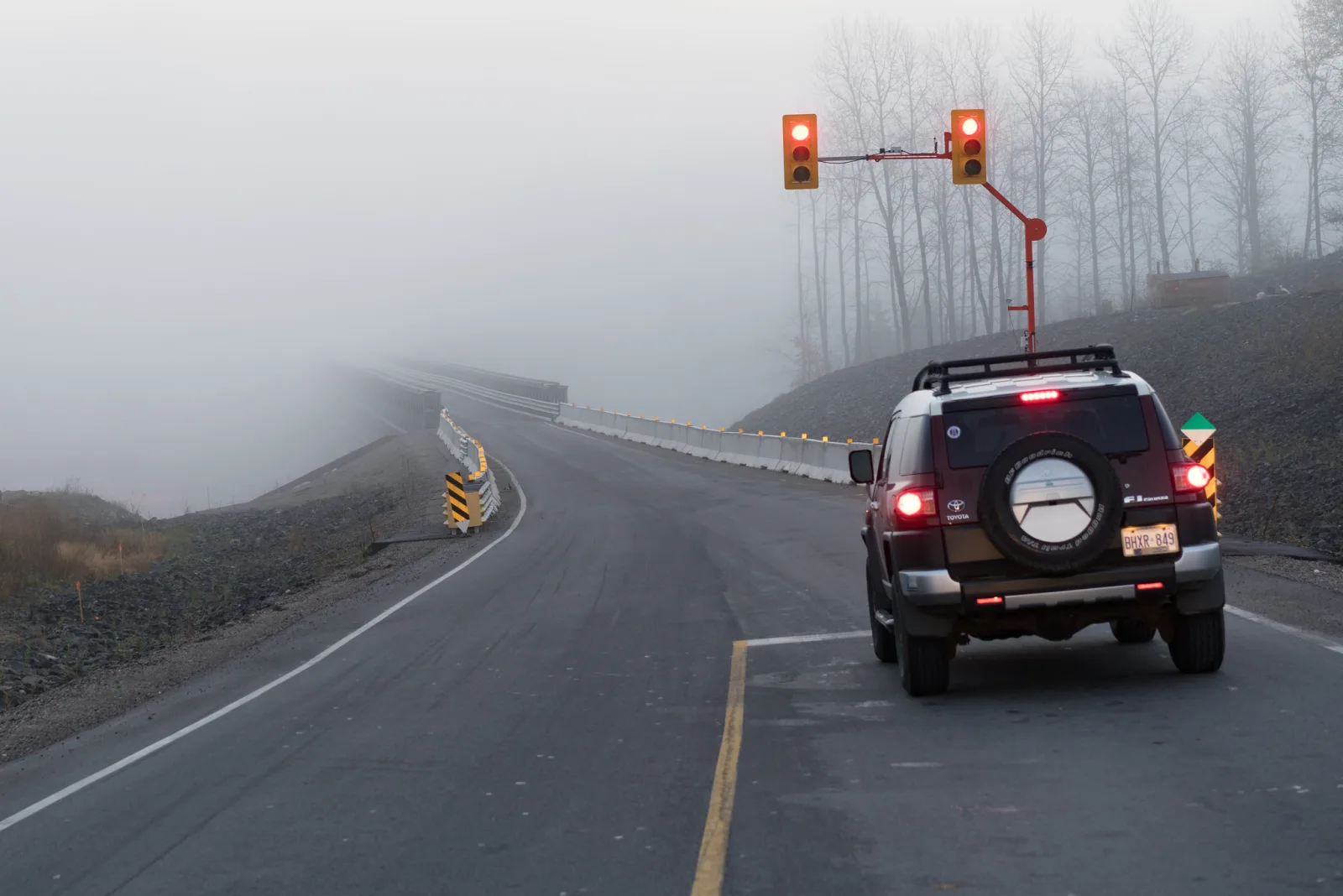 A dark SUV on a two-lane road beneath red traffic signals, with the road ahead curving away into thick fog and bare trees fading to grey on the hillside
