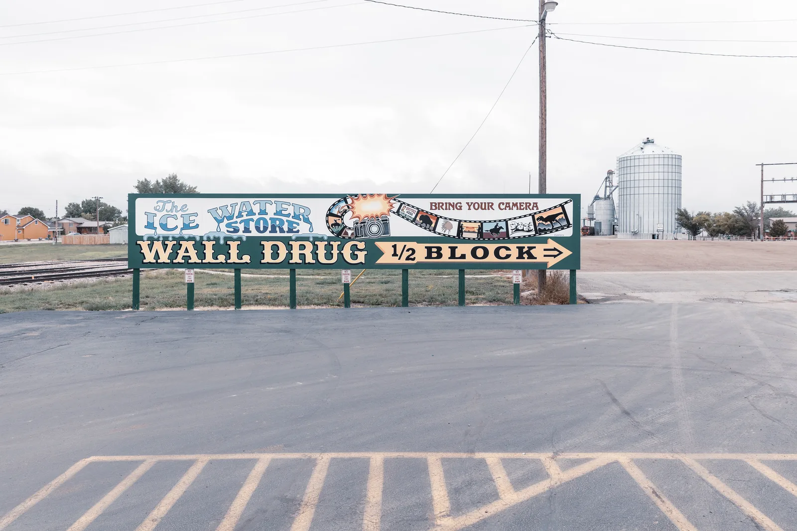 A long painted roadside billboard with large lettering standing at the far side of an empty car park under a pale overcast sky, with a grain silo behind it