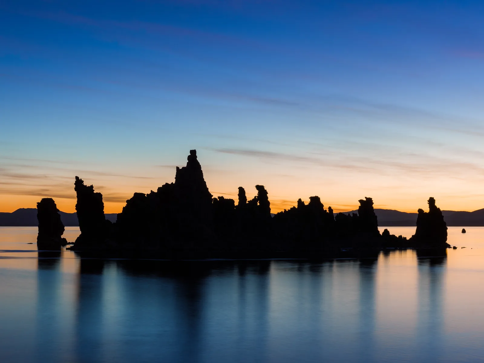 Jagged rock towers in silhouette on a still lake at dusk, with an orange band of light on the horizon fading up into deep blue sky