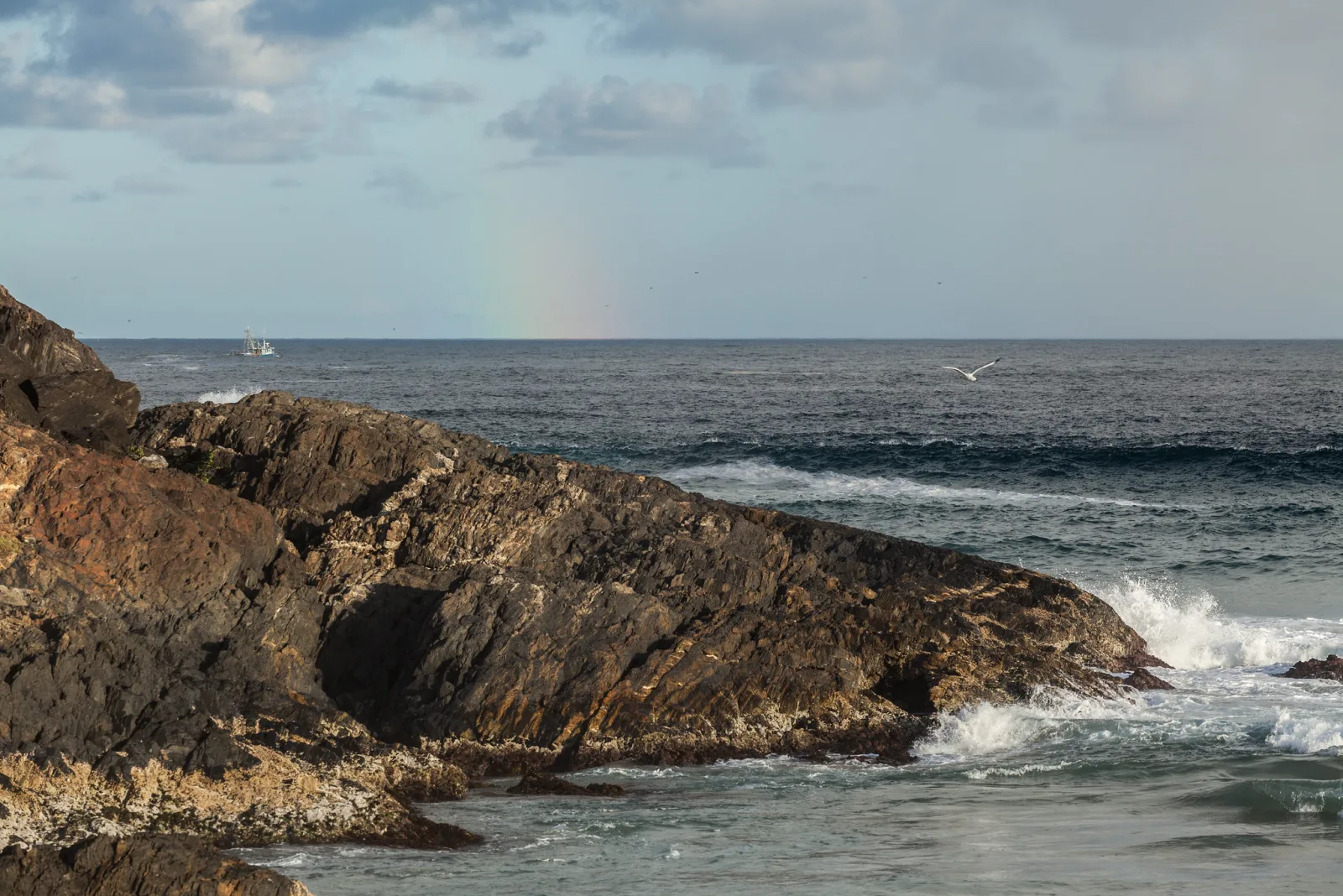 A dark rocky outcrop in the foreground with waves breaking white around its tip, open sea beyond and a faint rainbow over the horizon