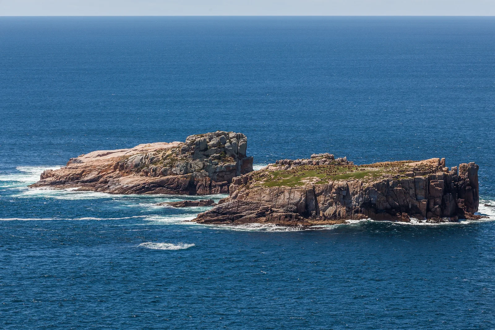 Two low rocky islets with white surf around them in deep blue sea, with a straight sea horizon and a band of pale sky across the top of the frame