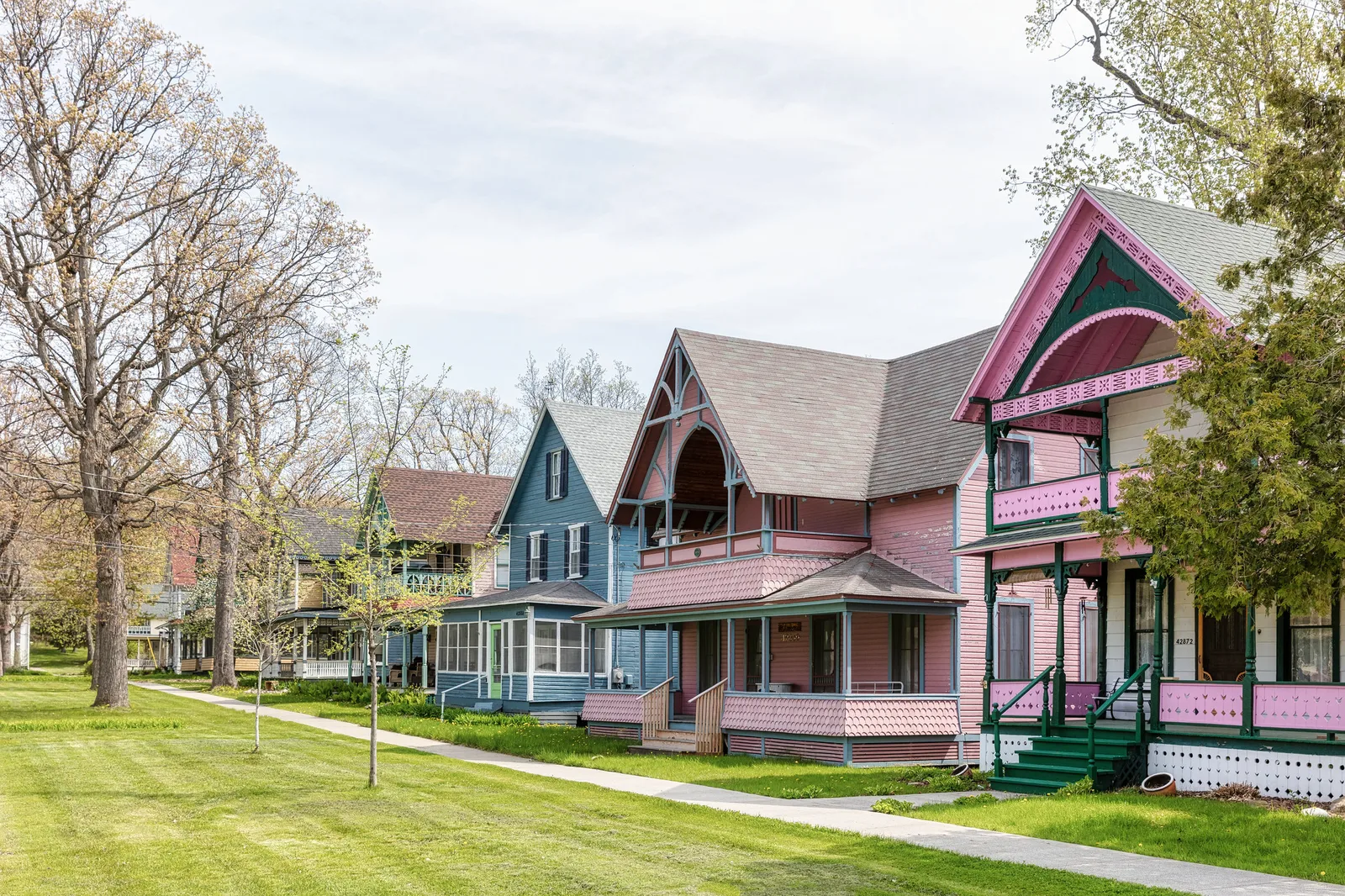 A row of pink, blue and green wooden houses with porches and decorative trim, seen at an angle across a mown lawn under bare spring trees