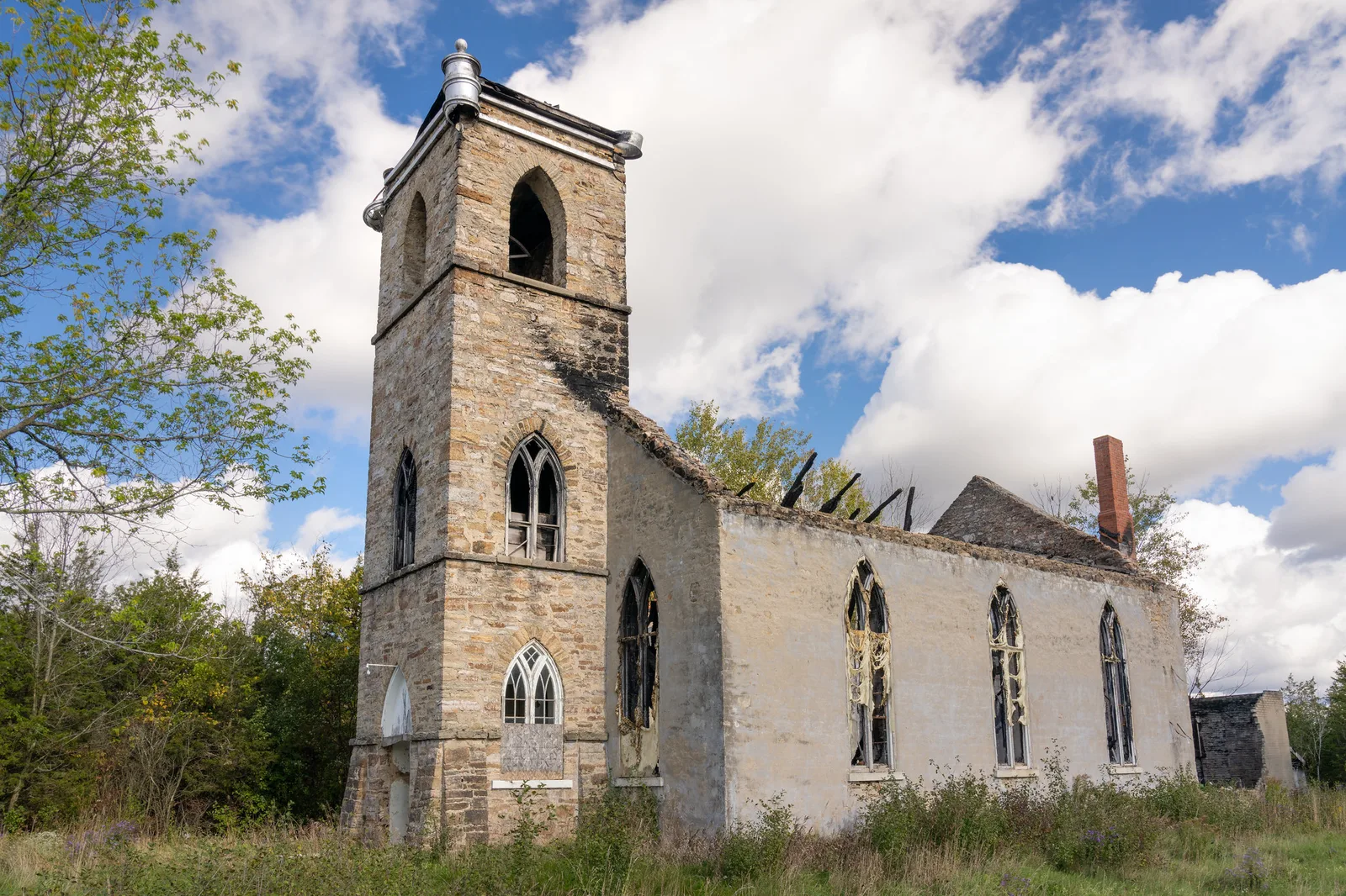 A roofless stone church with a square tower and empty arched windows, seen from a low corner angle under a blue sky with white clouds