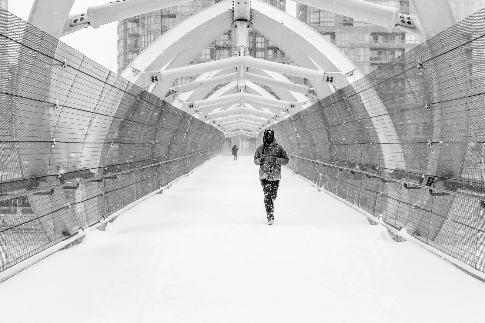 Black and white view down a snow-covered glass-walled pedestrian bridge with a lone runner in dark clothes in the middle of the walkway