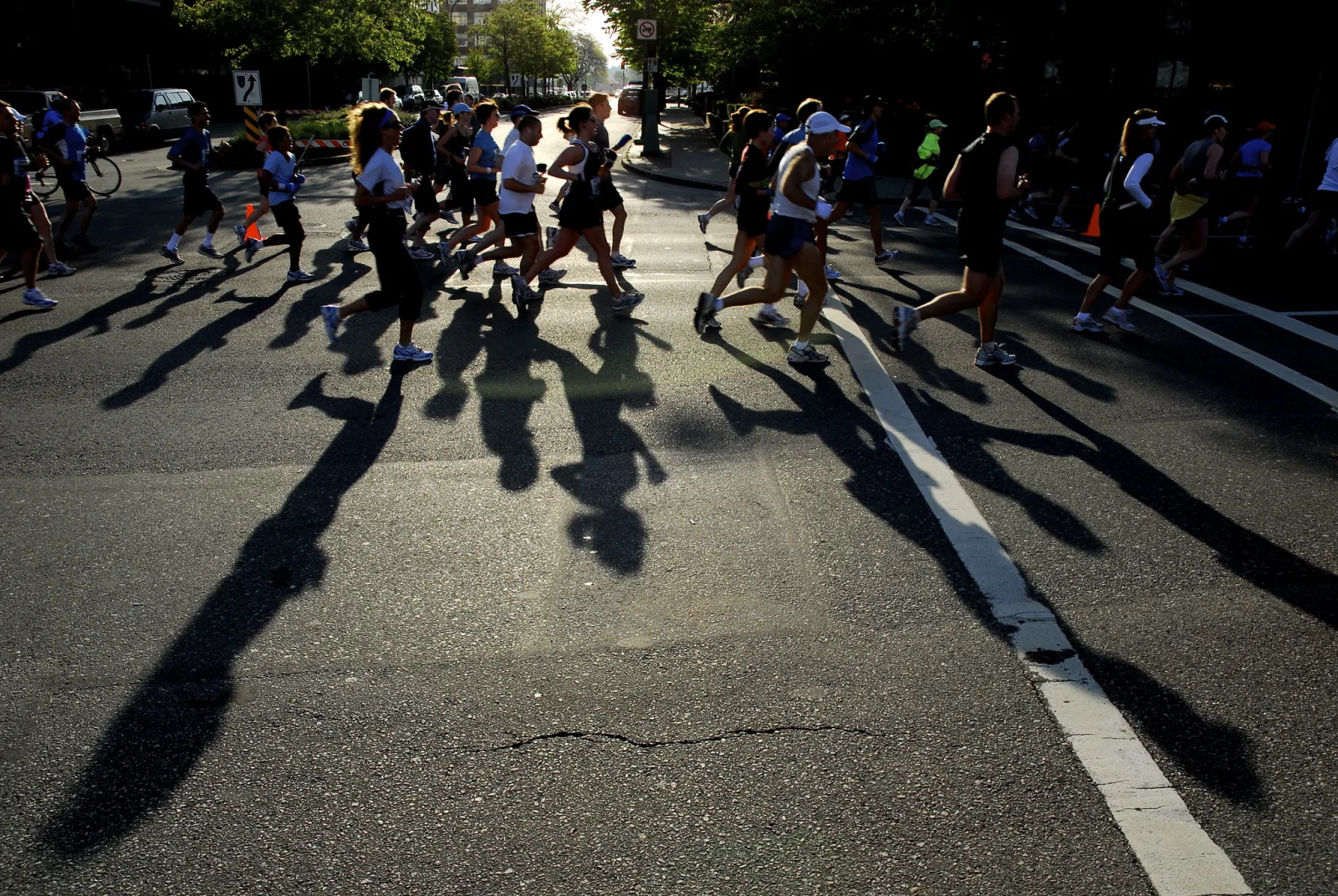 A crowd of runners crossing a sunlit road, their long black shadows stretching across the asphalt toward the camera