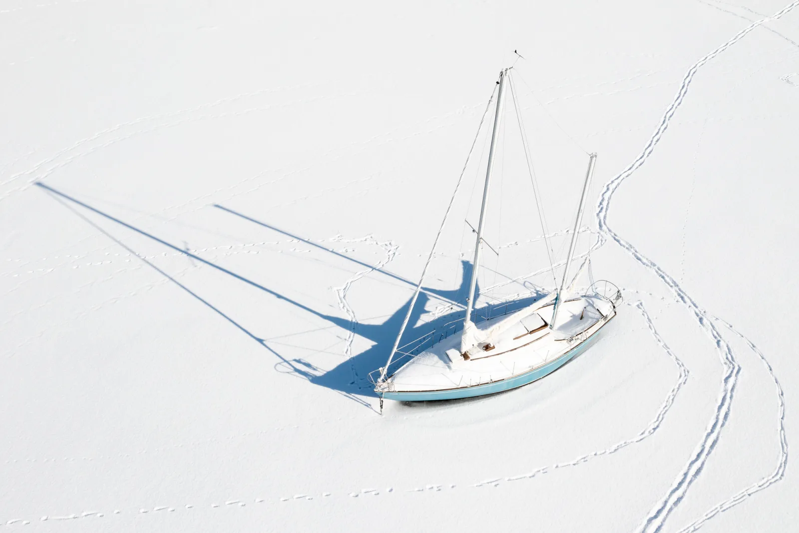 Aerial view of a sailboat frozen into white snow and ice, its masts and rigging casting long thin shadows across the snow, footprints curving around it