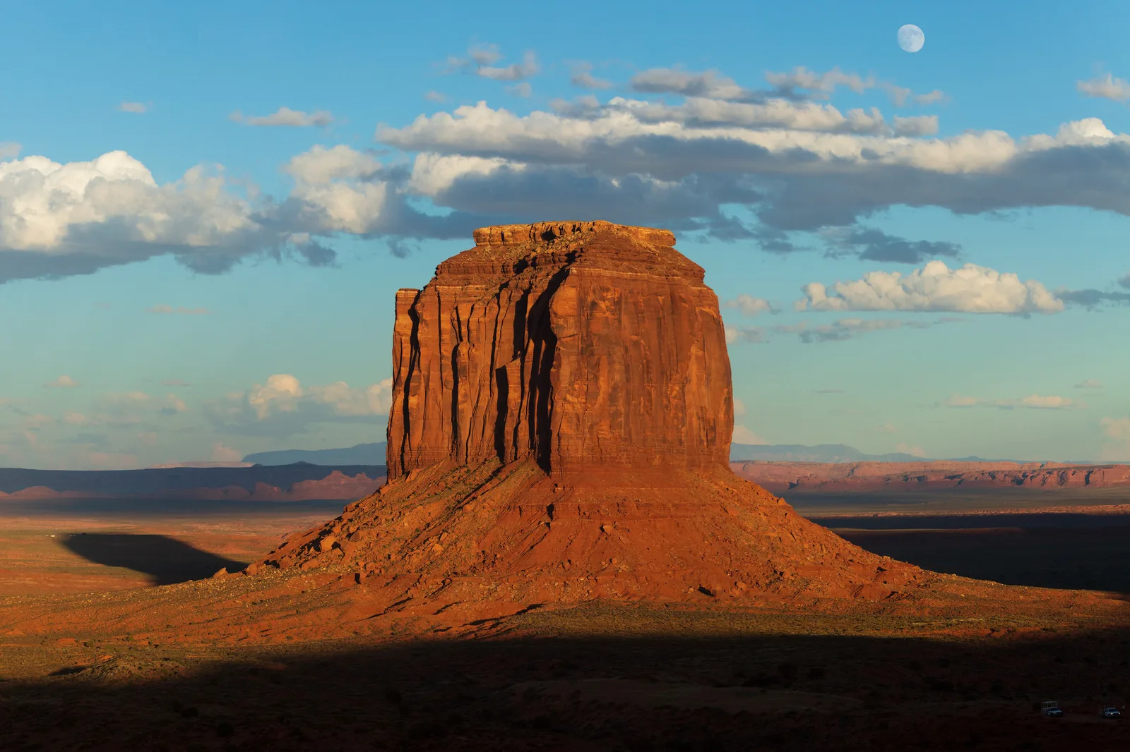 A lone sandstone butte lit orange by low sun on a desert plain, with a band of shadow across the foreground and a pale moon in a blue sky with scattered clouds