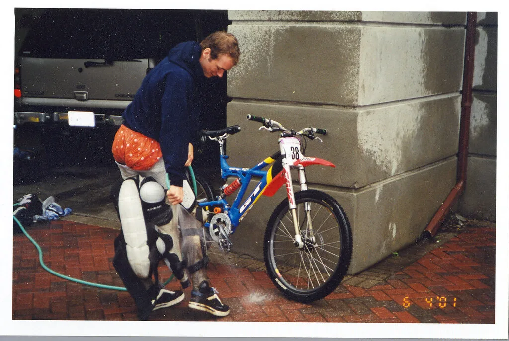 Scan of an old colour snapshot print, with its white border visible, showing a person hosing down a dirt bike beside a stone wall; orange date digits are printed in the lower right corner