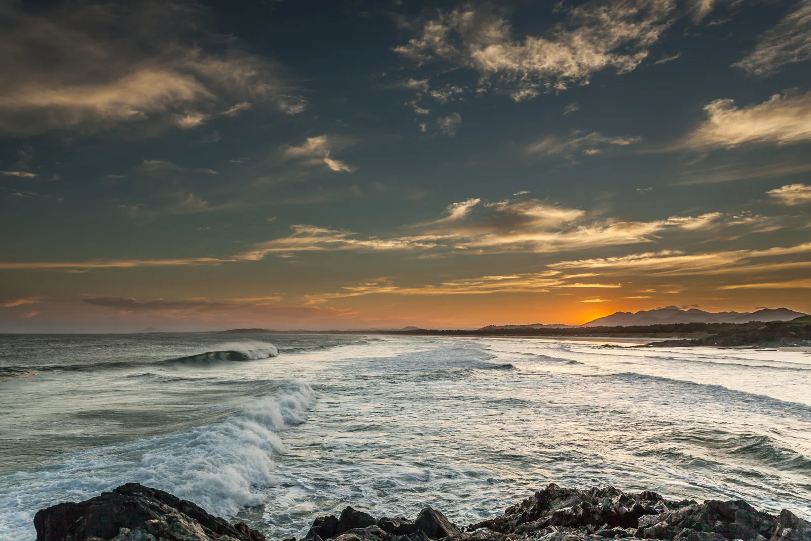 Waves rolling onto a rocky shore at sunset, dark rocks in the foreground, white foam across the water and orange light on the horizon under scattered clouds