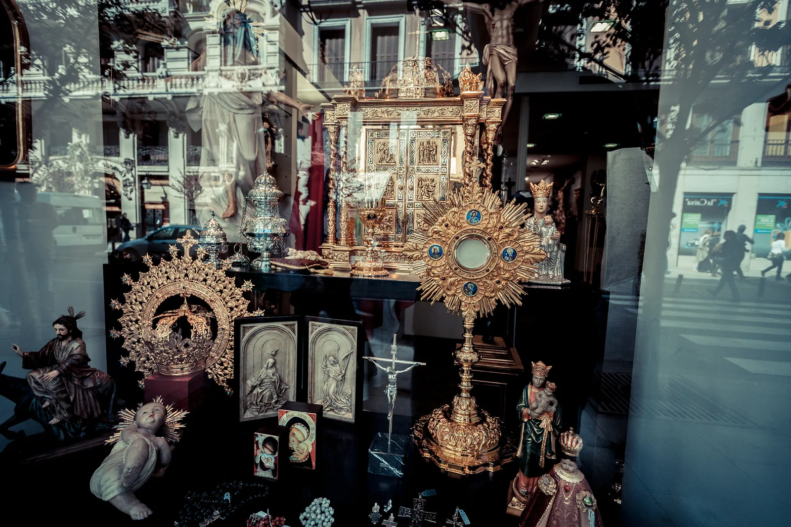 A shop window display of gilded religious objects and figurines, with the buildings, trees and pedestrians of the street behind the camera reflected across the glass