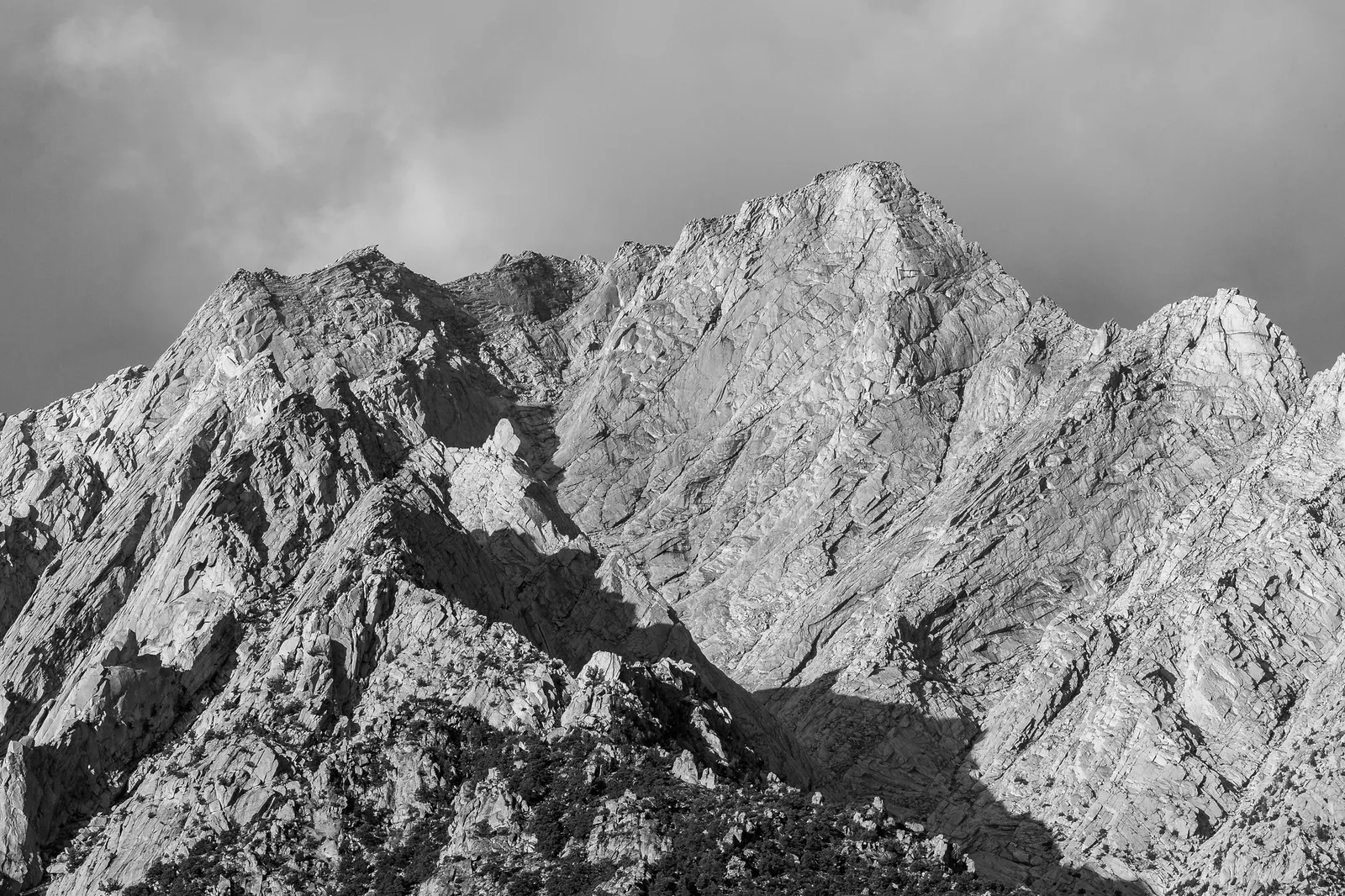 Black and white view of a jagged, cracked granite peak under grey cloud, with one face in sunlight and the gullies in deep shadow
