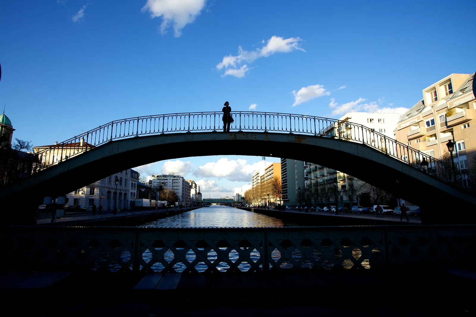 A single person in silhouette standing at the top of an arched iron footbridge over a canal, against a deep blue sky with scattered white clouds