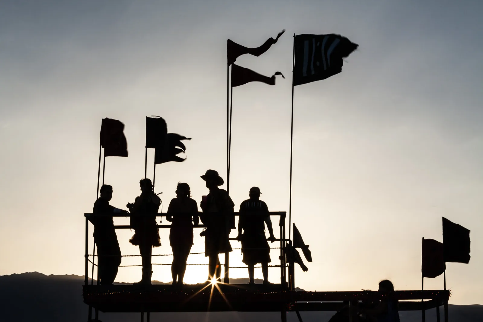 Five people in silhouette standing at the railing of a raised platform with black flags, against a hazy evening sky with the sun behind them