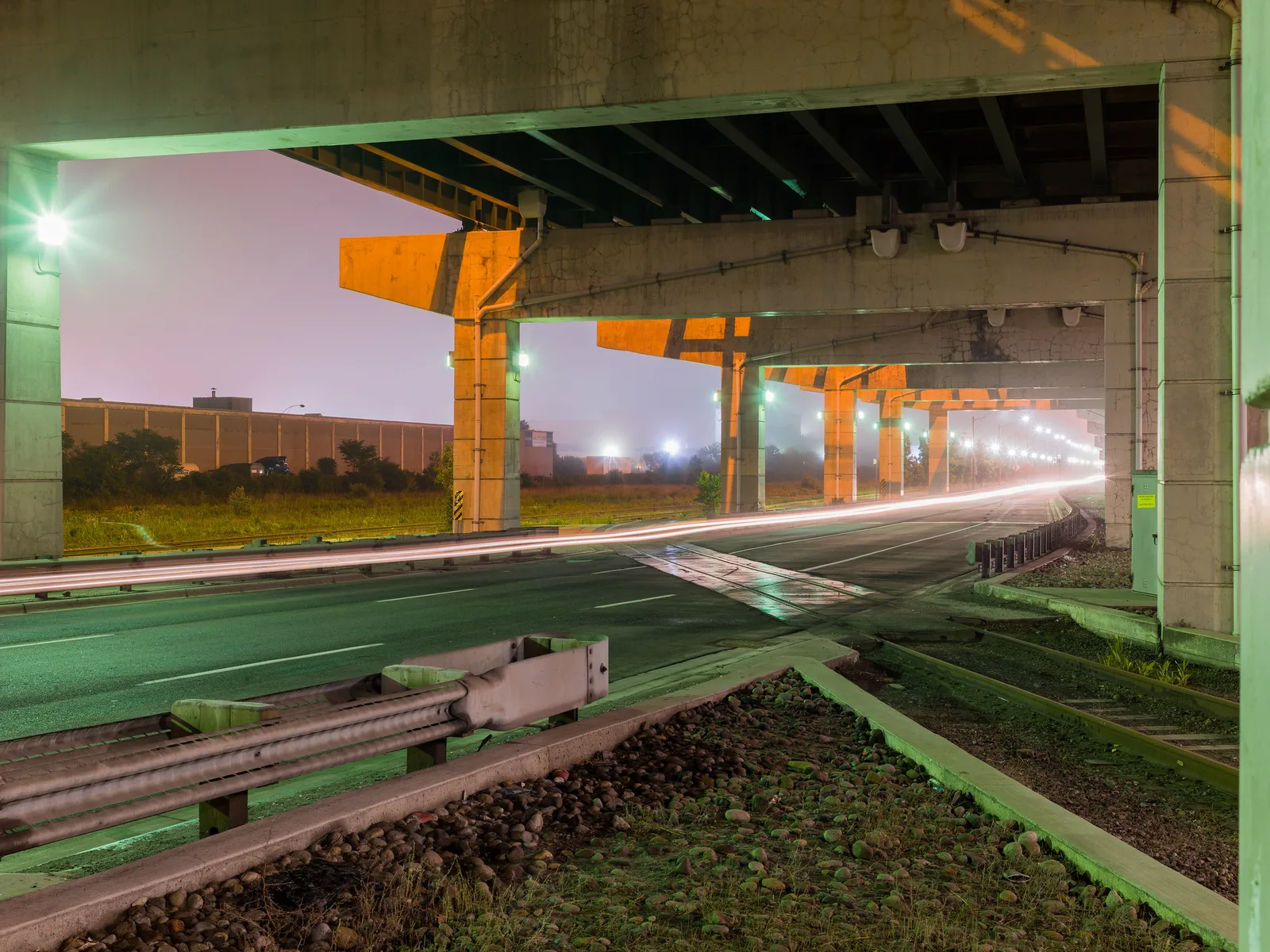 A road passing beneath a concrete elevated expressway at night, with one continuous white streak of light running along the lane and orange-lit pillars receding into the distance