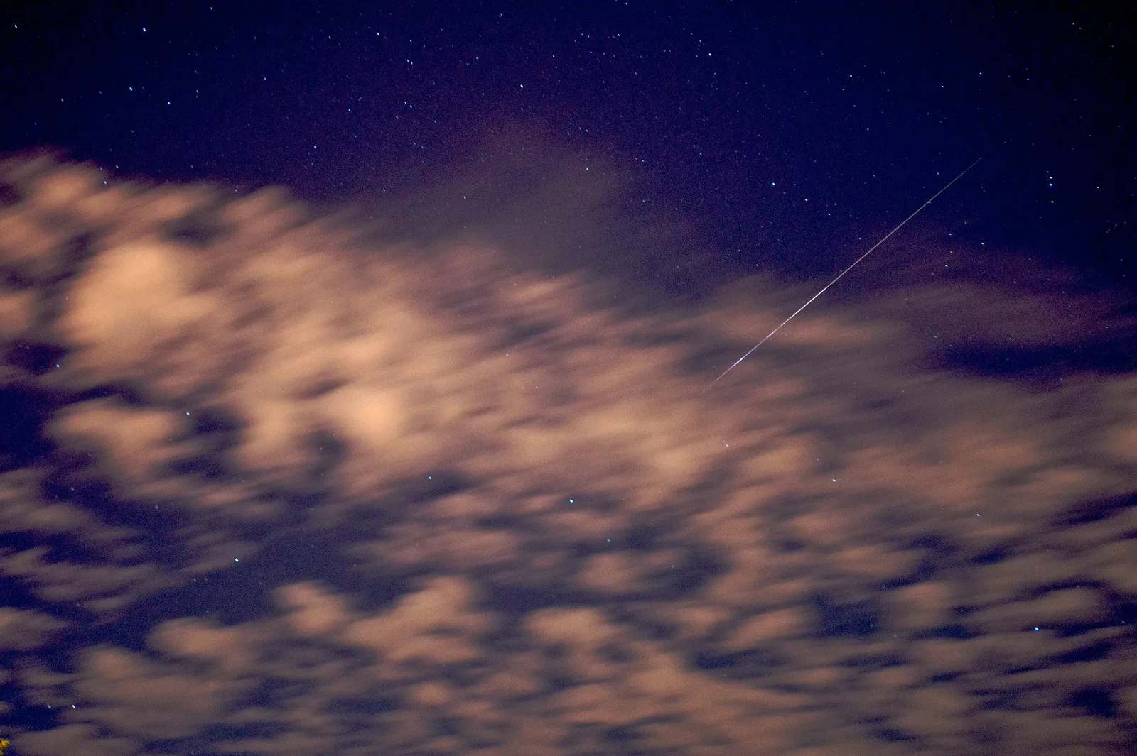 A single straight meteor streak crossing a deep blue starry sky above a bank of blurred, softly lit clouds