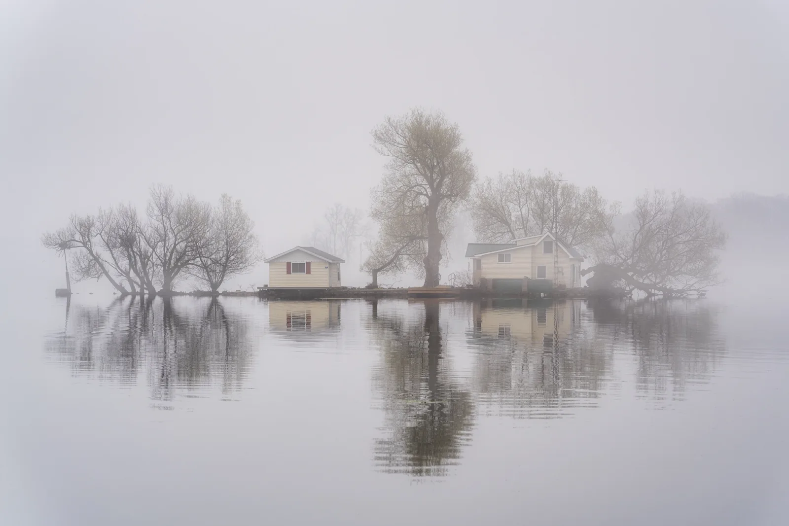 Two small cottages and bare trees on a tiny island, softened by thick fog and mirrored in still grey water