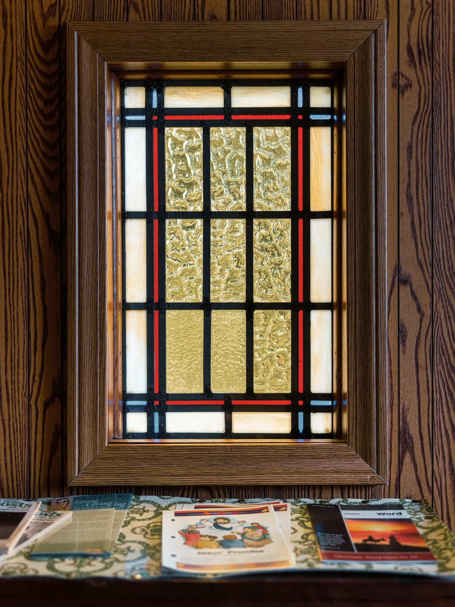 A small rectangular stained glass window with red and blue leaded lines and textured yellow glass, set in a dark wooden frame above a table of leaflets