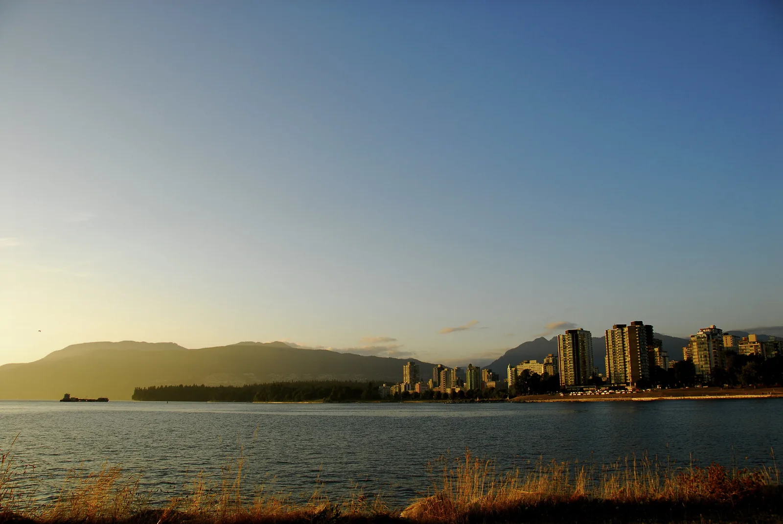 A clear evening sky fading smoothly from pale yellow near the sun to deep blue, above a bay with distant apartment towers and hills