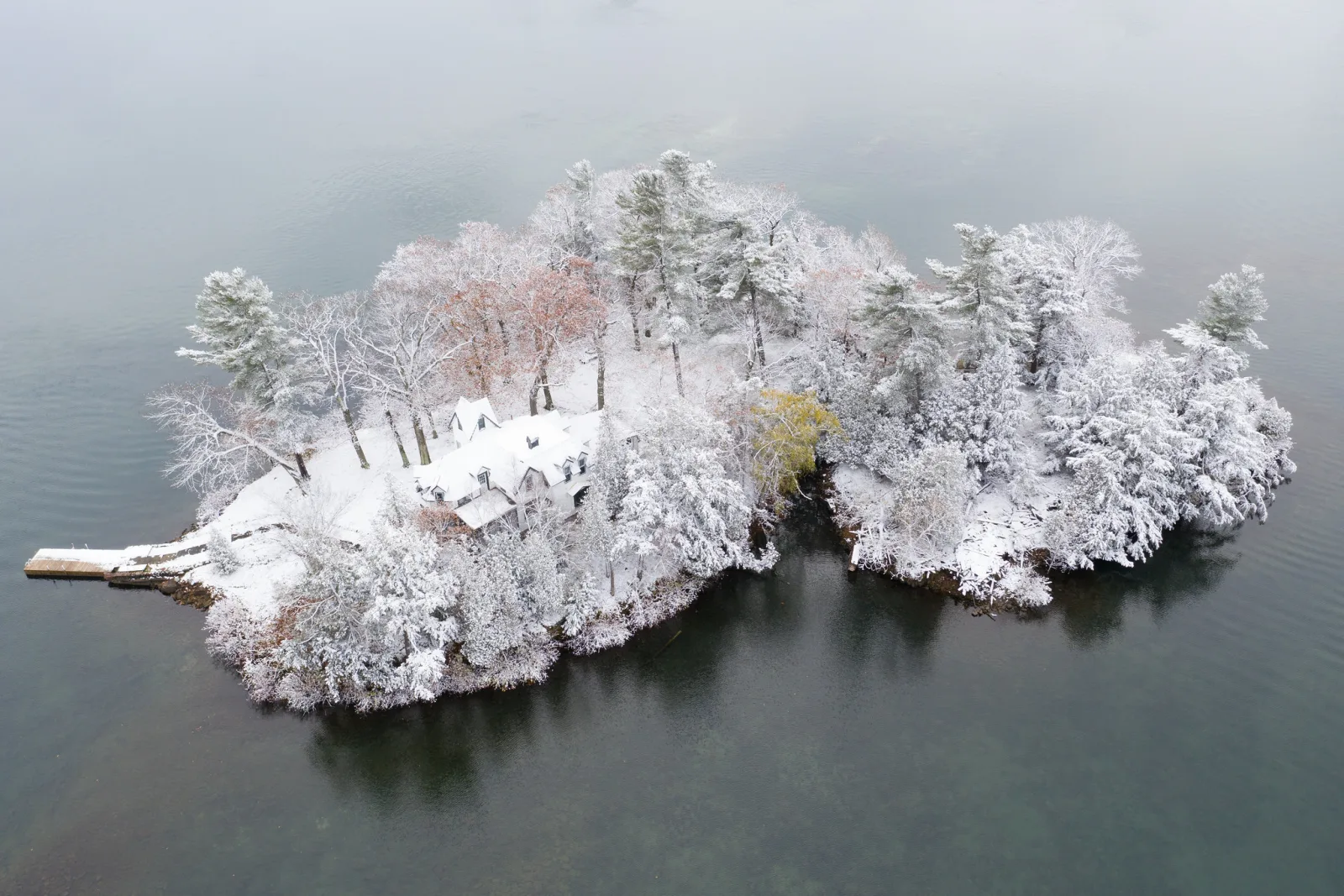 Aerial view of a small wooded island covered in fresh snow, with a cottage and dock among white trees and dark grey water around it