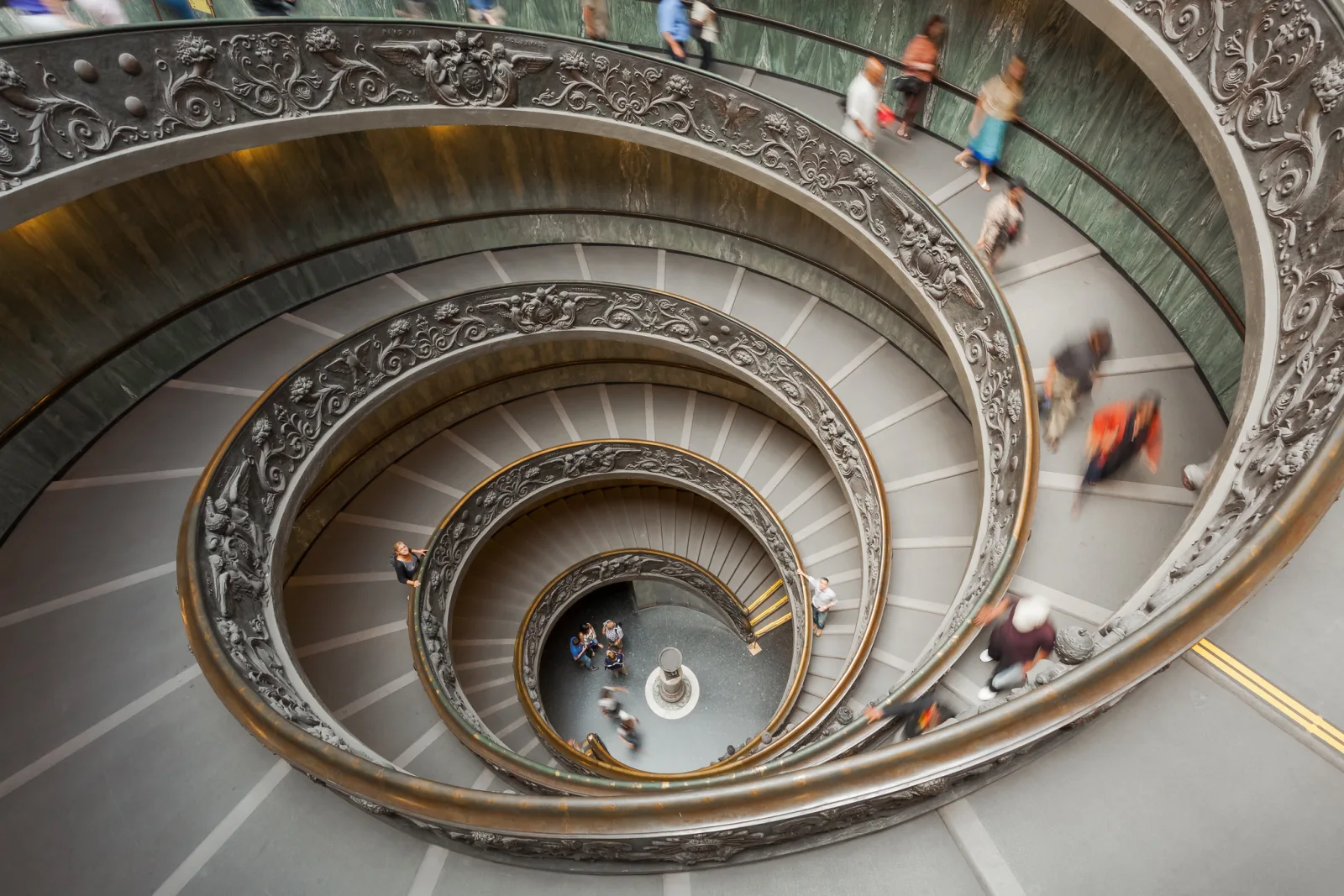 Looking down an ornate spiral staircase from the top, with visitors walking down the ramp in soft motion blur and the spiral narrowing to a small floor far below