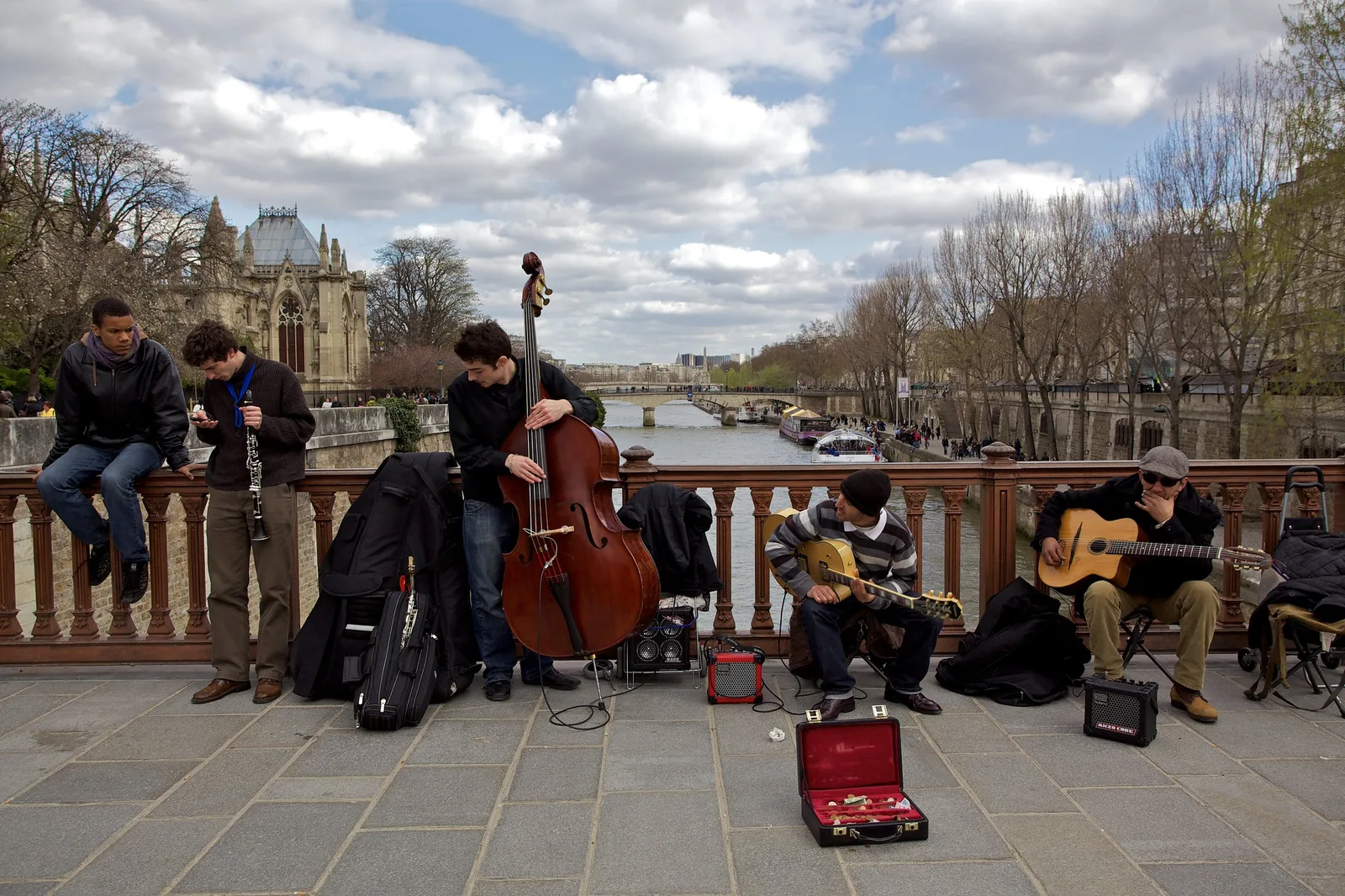 A group of young street musicians with a double bass, clarinet and guitars playing against a bridge railing, with an open instrument case on the pavement and a river behind