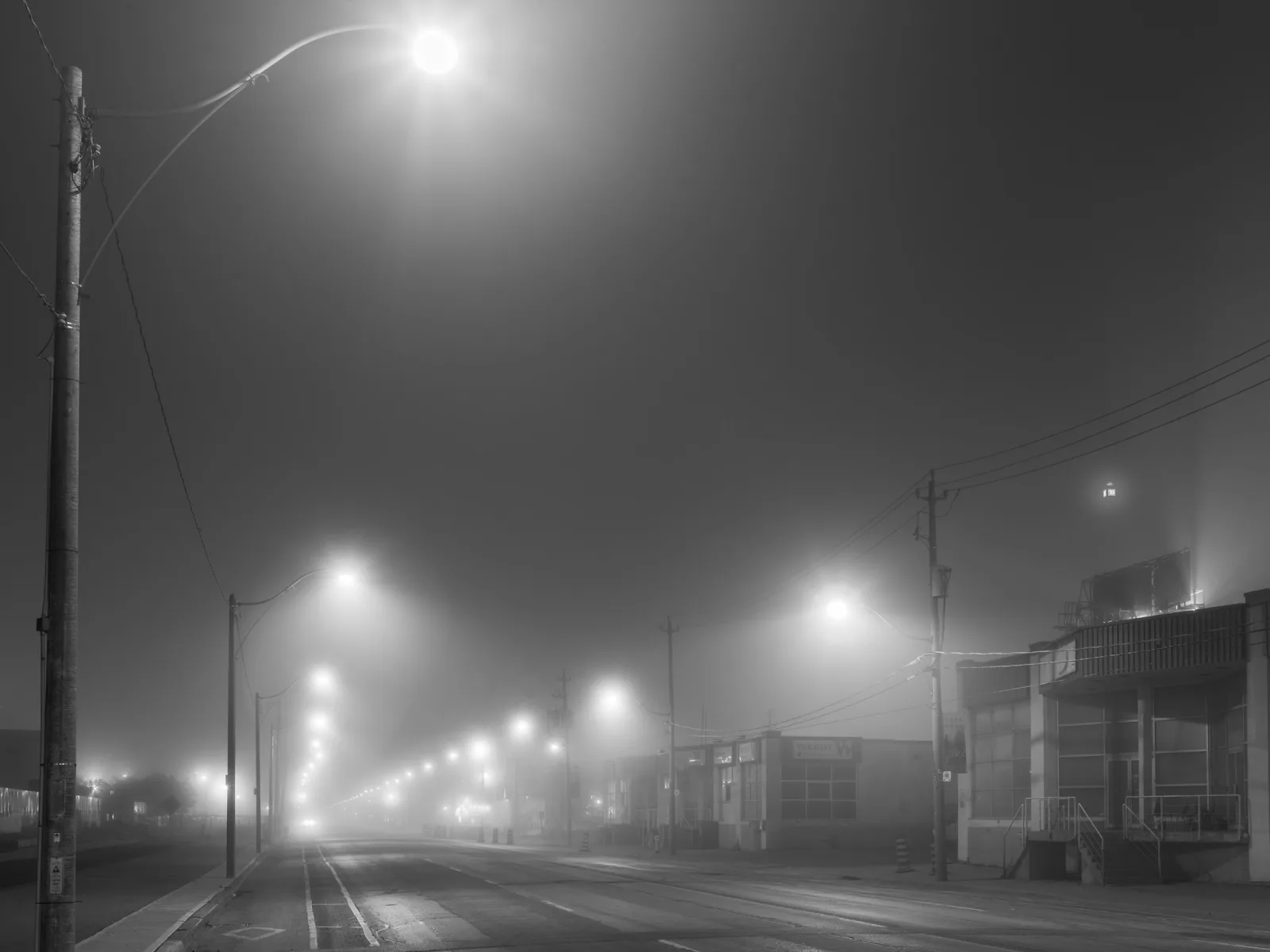 Black and white night view down a straight street with a line of street lights glowing in fog, each light fainter and softer than the one before