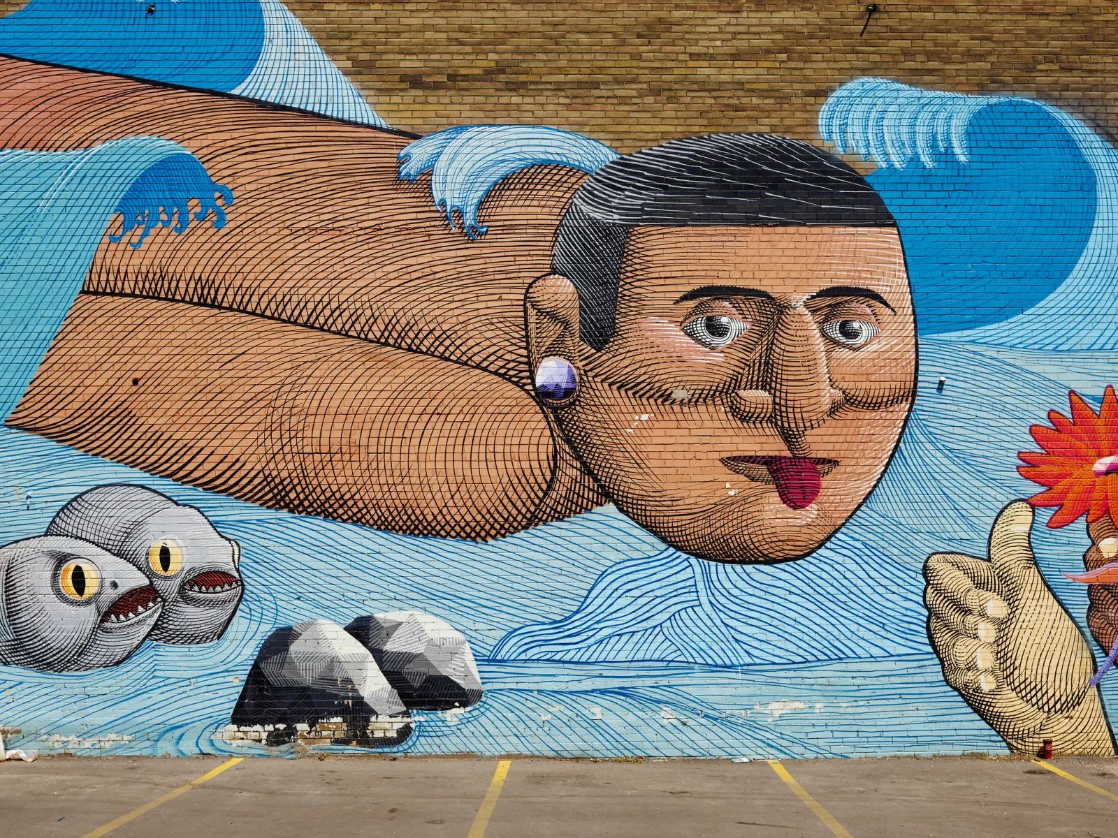 A large painted mural of a man flying horizontally over stylized blue waves with fish and rocks, on a brick wall above a parking lot