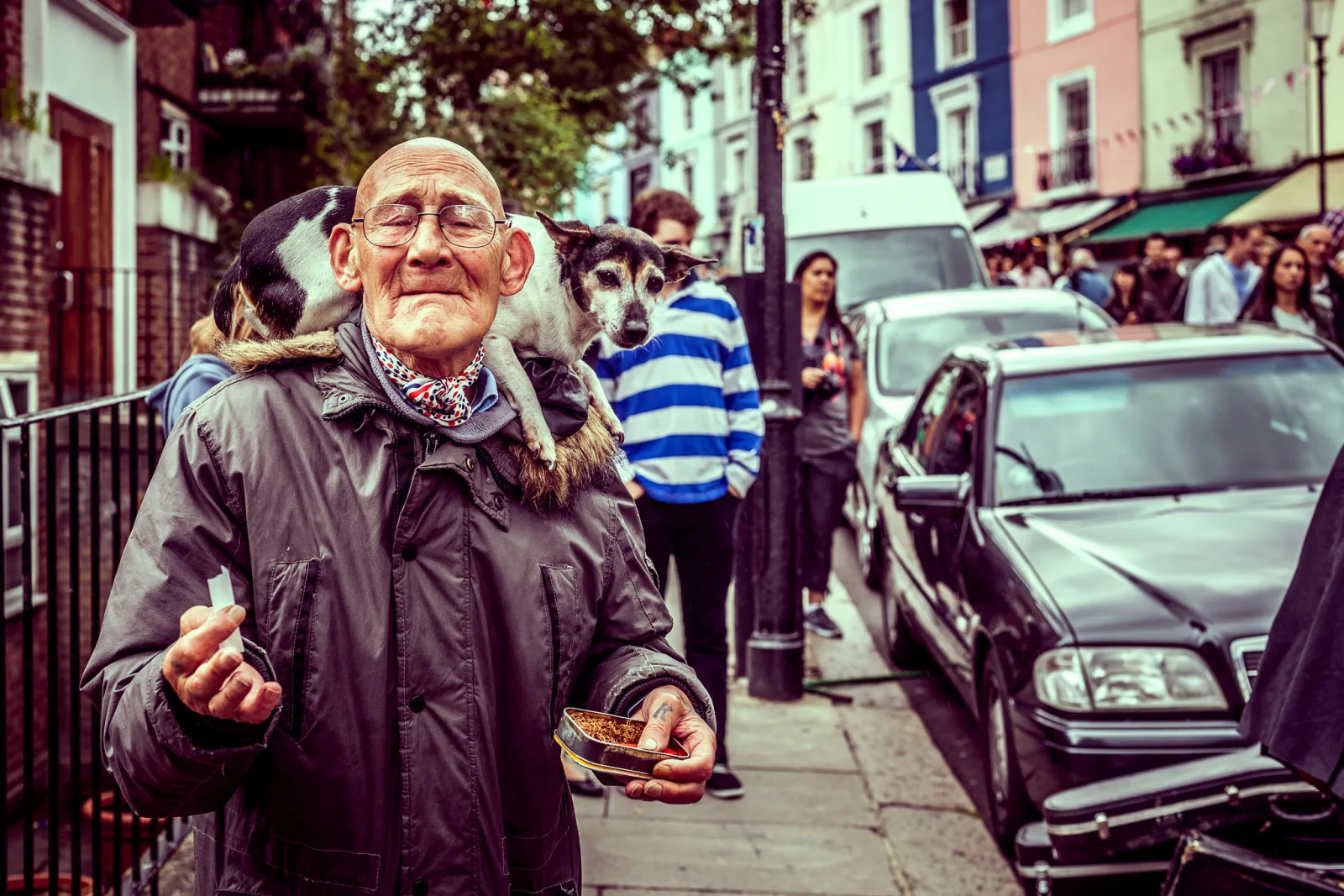 An older man in glasses and a dark coat standing on a busy city sidewalk with a small dog perched on his shoulder, parked cars and pedestrians behind him