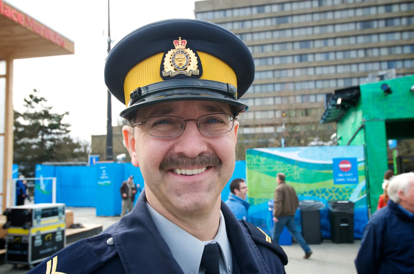 A smiling police officer in a peaked cap with a badge and glasses, framed from the shoulders up, with a street scene and temporary event structures behind him