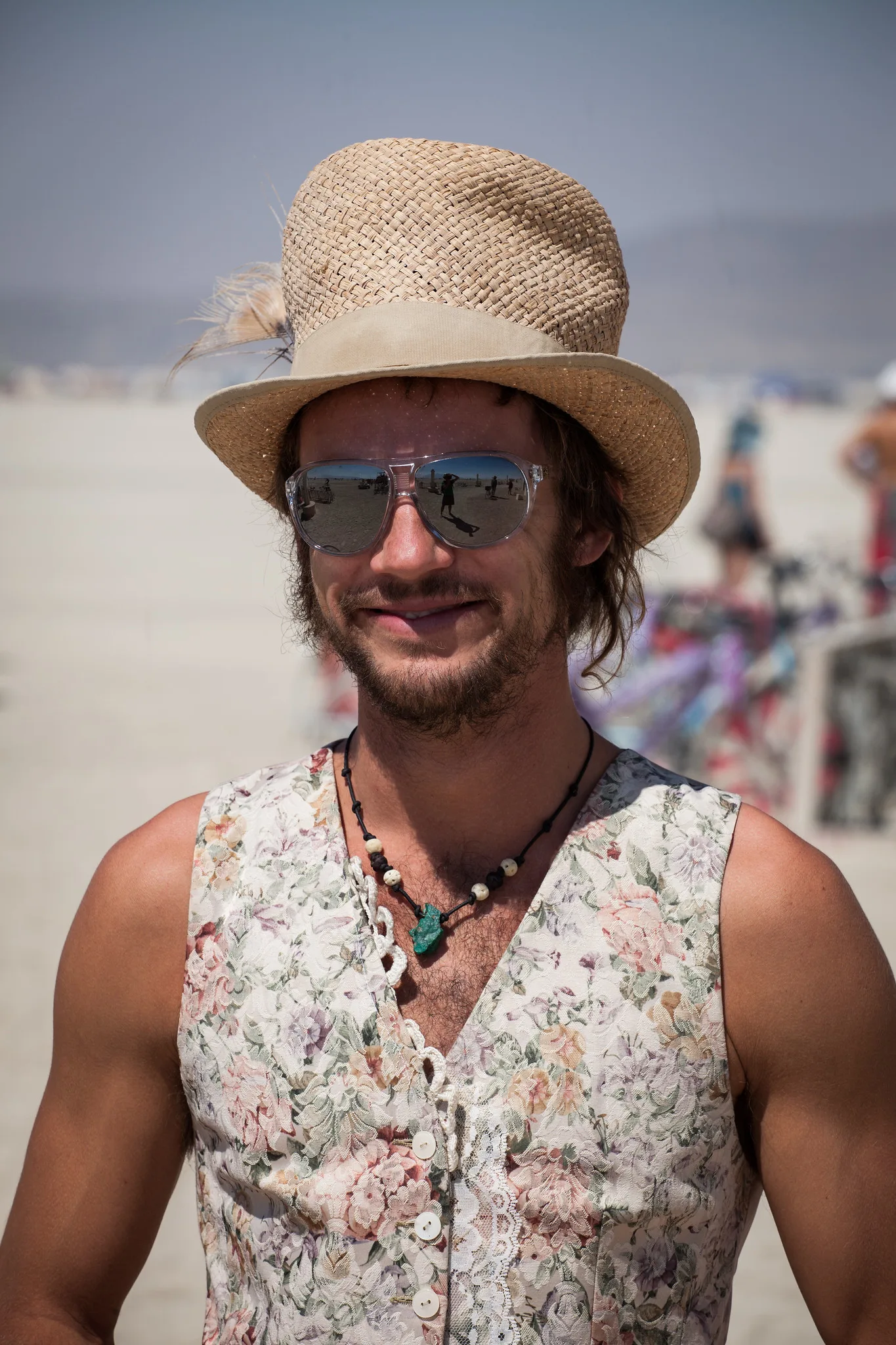 A smiling man in a straw hat, mirrored sunglasses and a floral sleeveless shirt, photographed from the chest up against a softly blurred bright background
