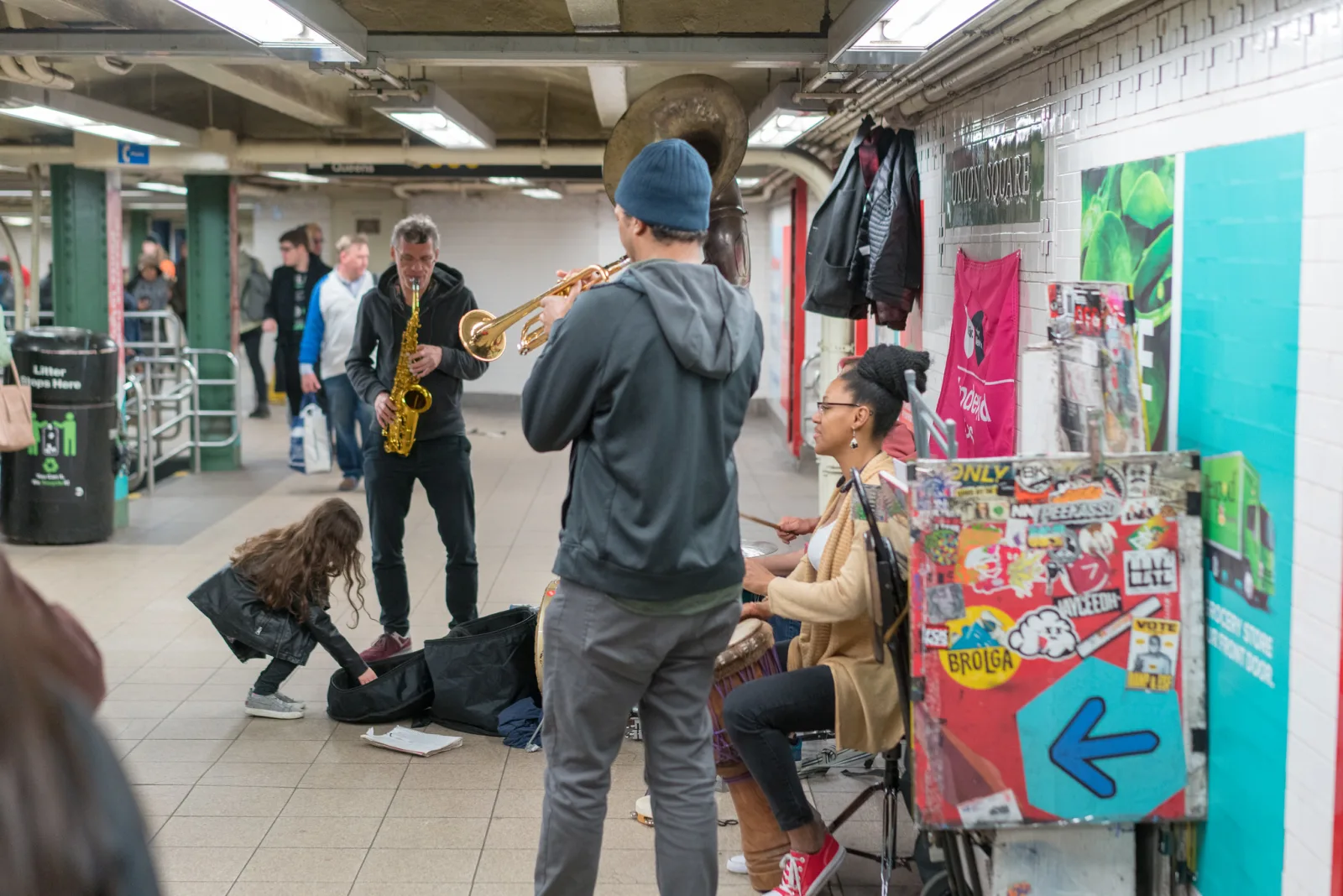 Street musicians playing saxophone, trumpet and keyboard on a subway platform under artificial station lighting, with a man bending over an open instrument case