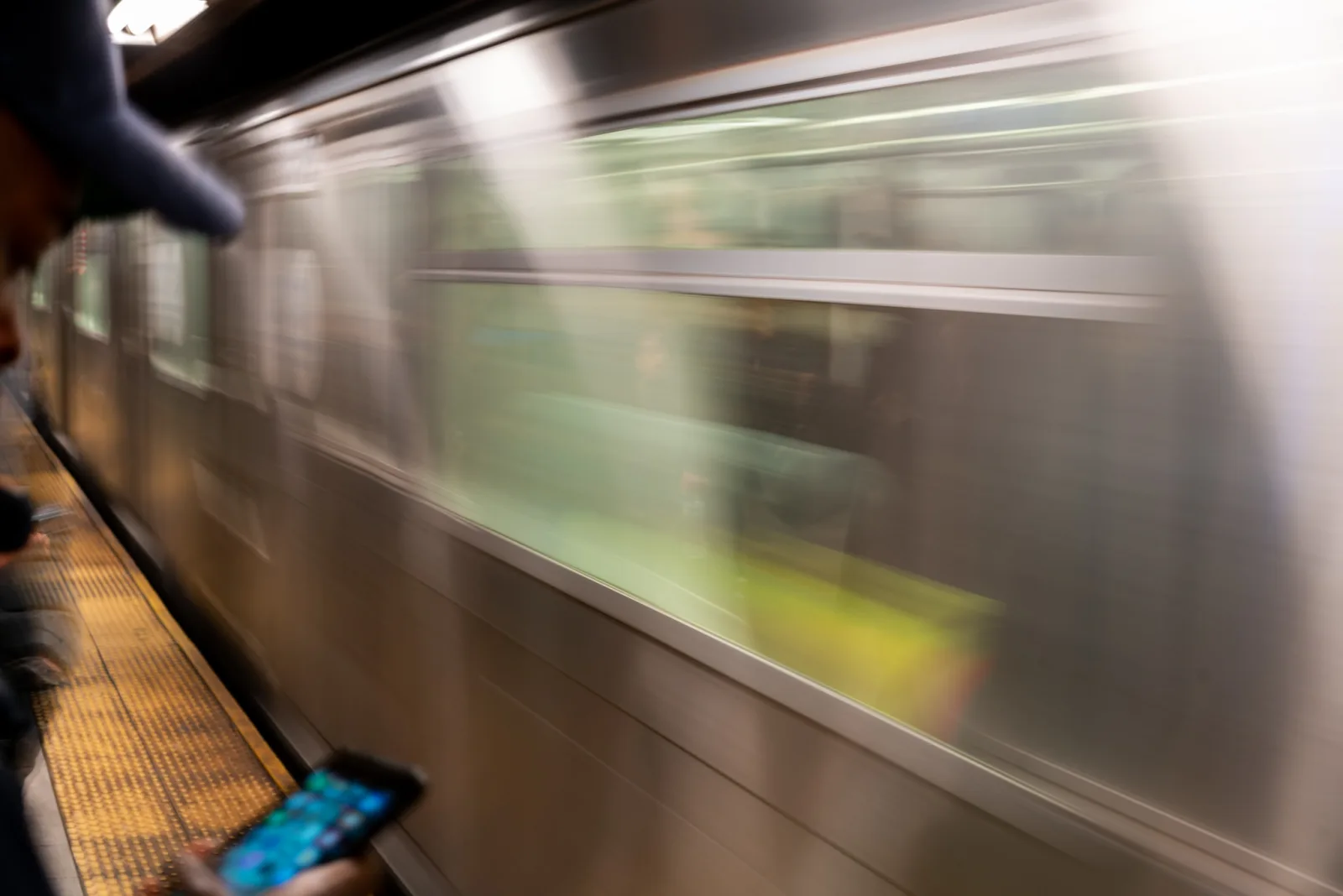 A subway train streaking past as a blur of green and silver, while a person on the platform at the left holds a lit phone that stays sharp