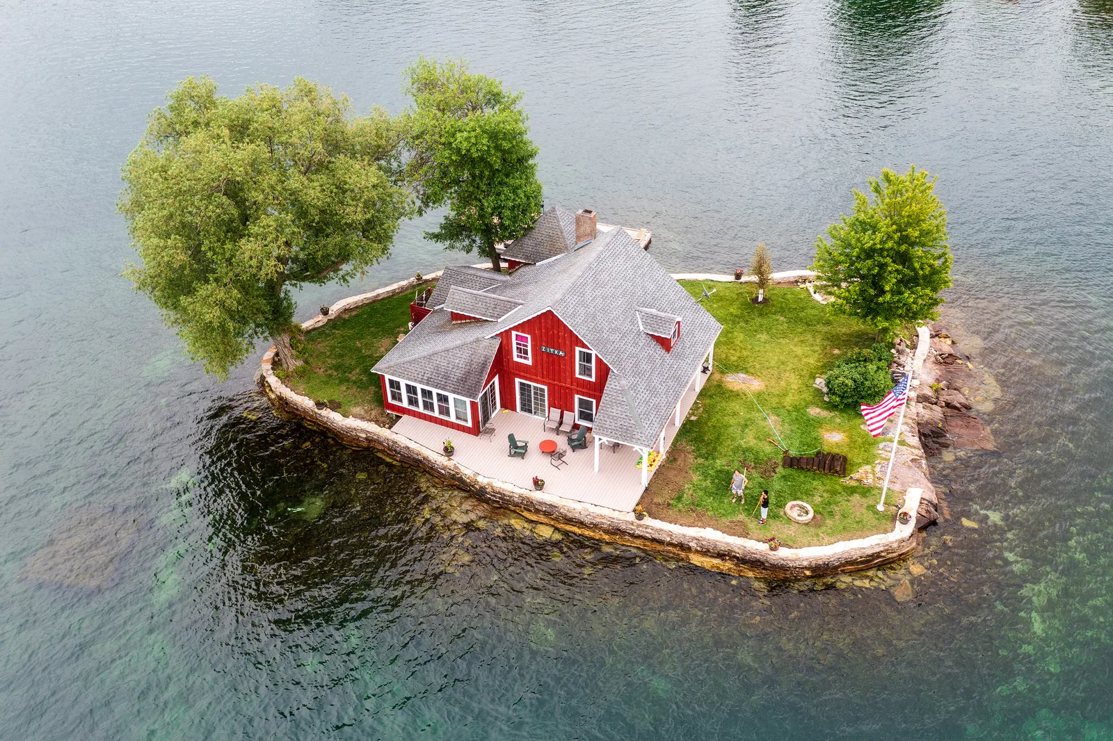 Aerial view of a small island in summer with a red cottage, green lawn and leafy trees, surrounded by calm green water
