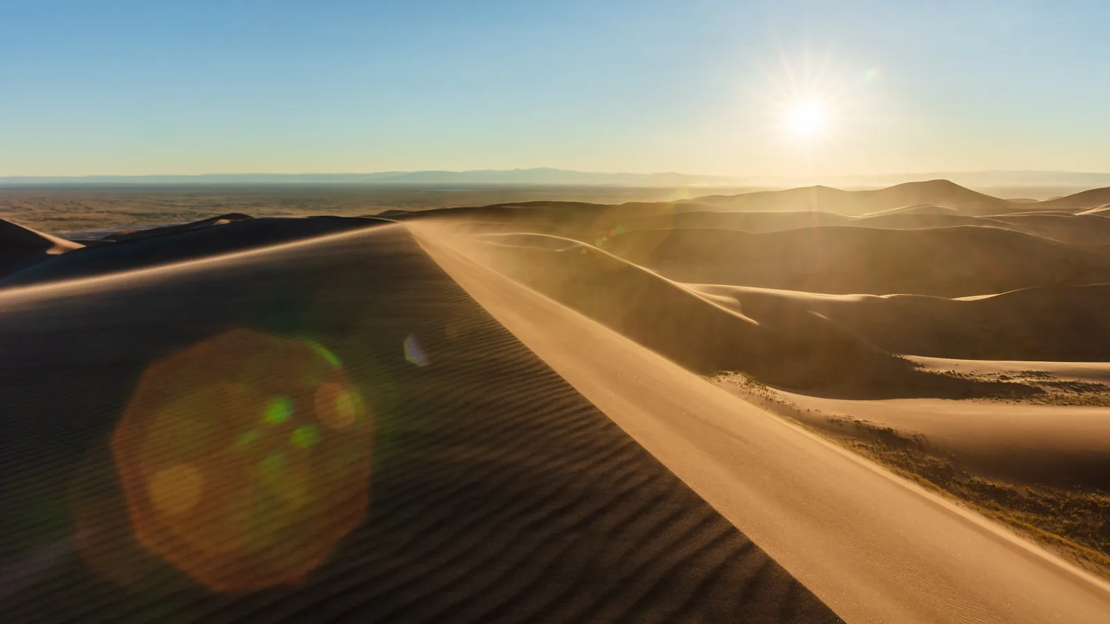 A sharp-crested sand dune running diagonally towards distant dunes, with the sun high in the frame and a large orange and green flare spot over the shadowed sand