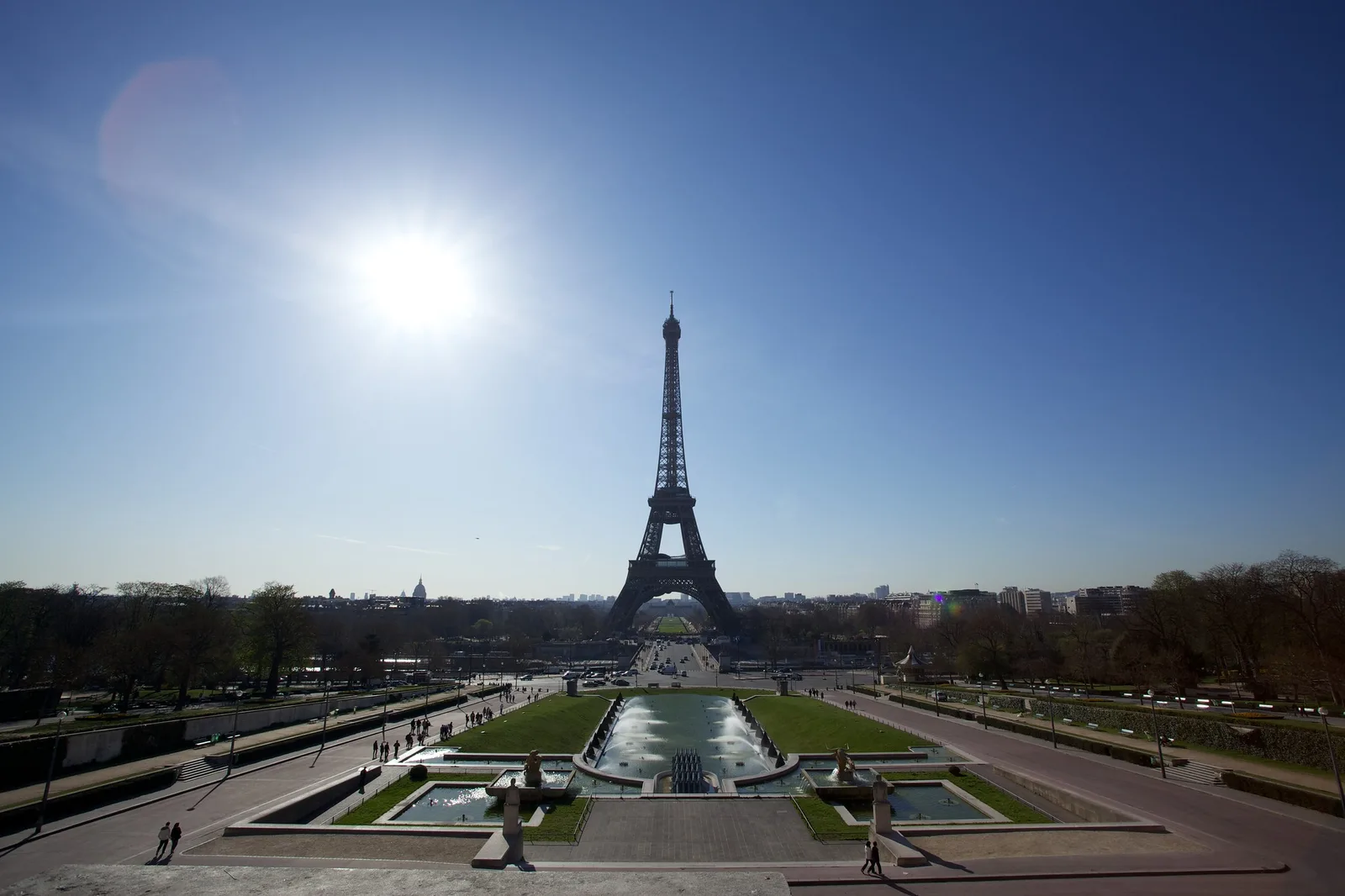 A wide view of the Eiffel Tower and the gardens in front of it with the sun in the upper left of a clear blue sky, a pale hazy flare patch floating beside the sun