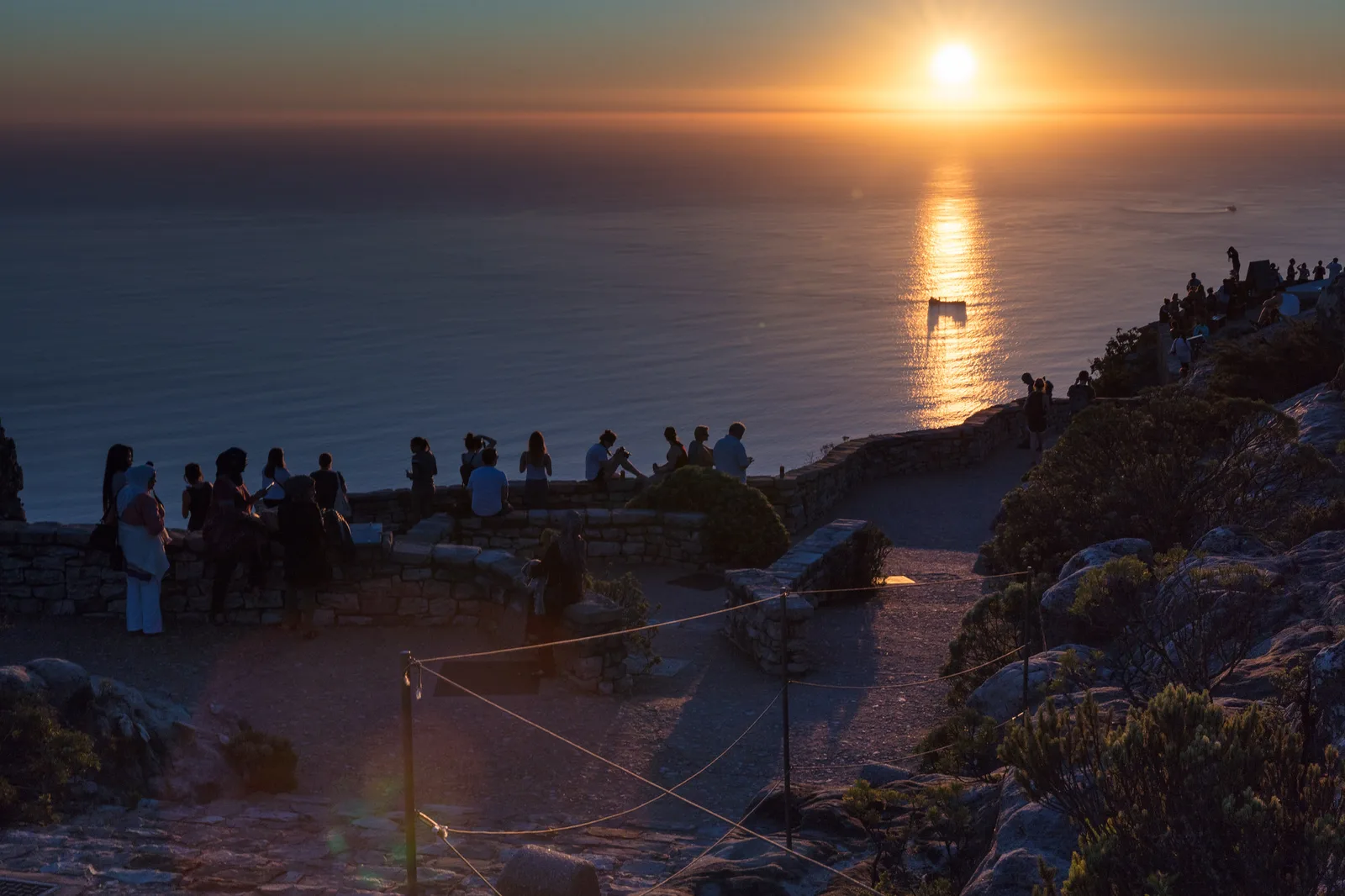 The sun low over the sea with a bright reflection on the water, and small groups of people gathered on a rocky viewpoint in the dim foreground