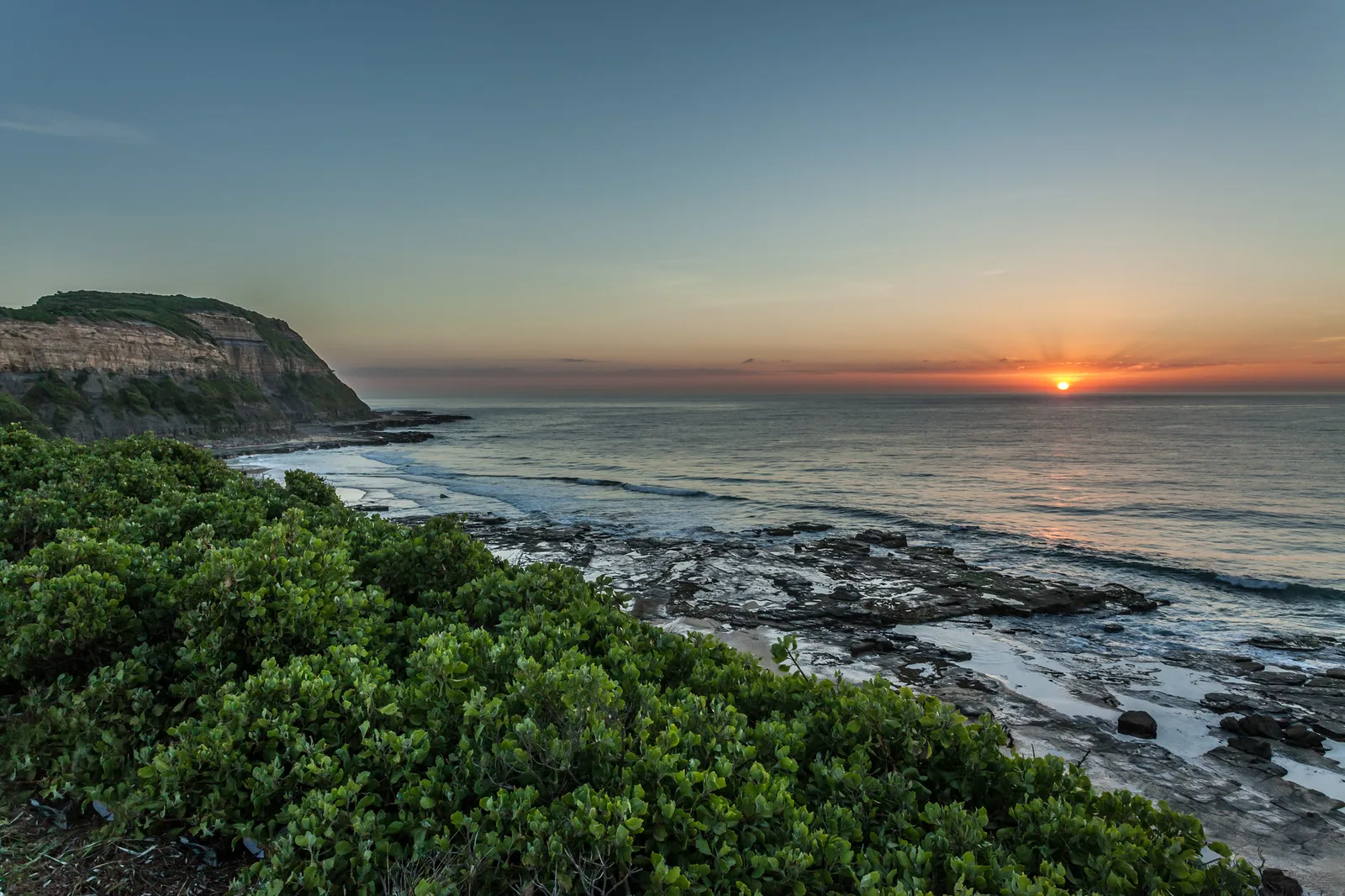 The sun sitting on the sea horizon under an orange and grey sky, seen from a headland with dense green shrubs in the foreground and a rocky shoreline below