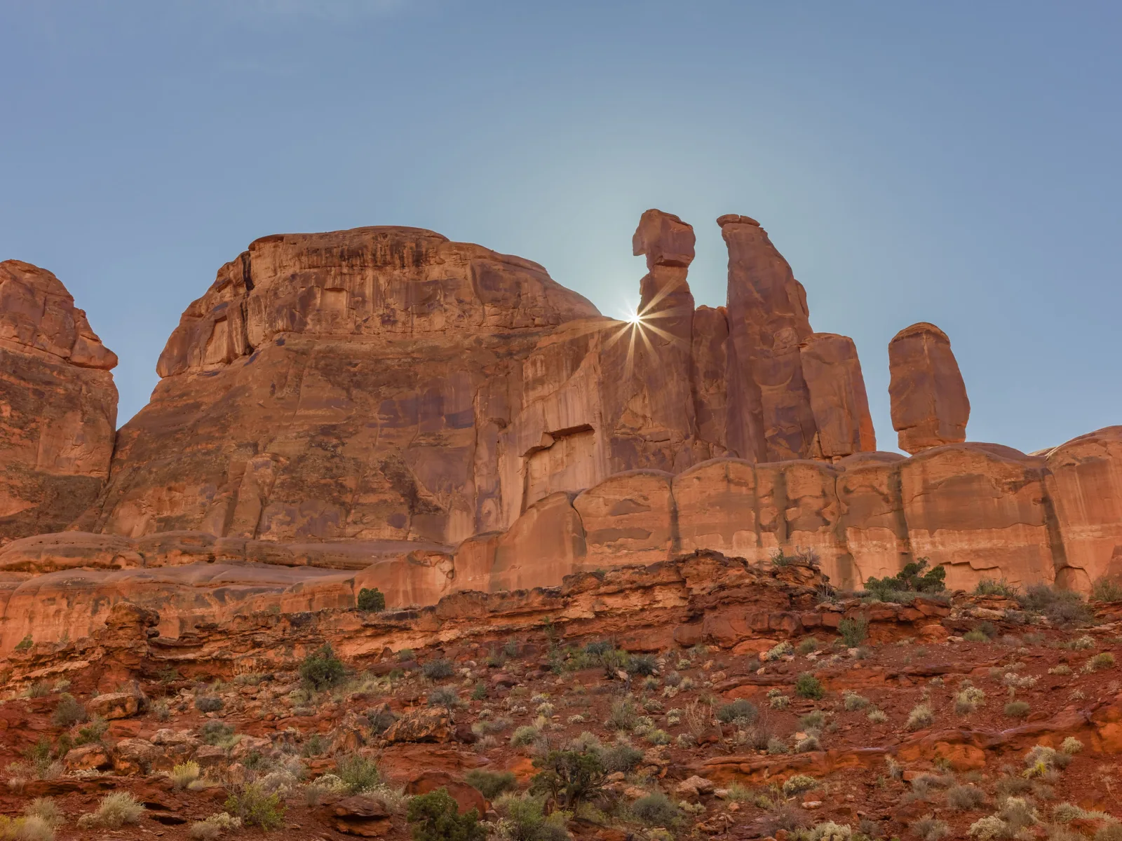 Red sandstone cliffs and spires under a clear blue sky, with the sun peeking from behind the edge of a spire as a small sharp sunstar