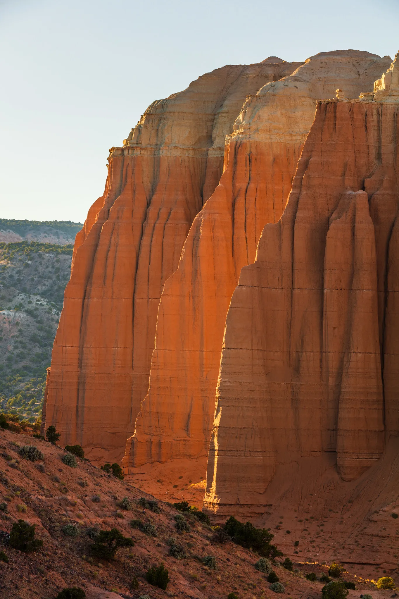 A tall red sandstone cliff with vertical ridges glowing in warm light, framed in a vertical format with a hillside below