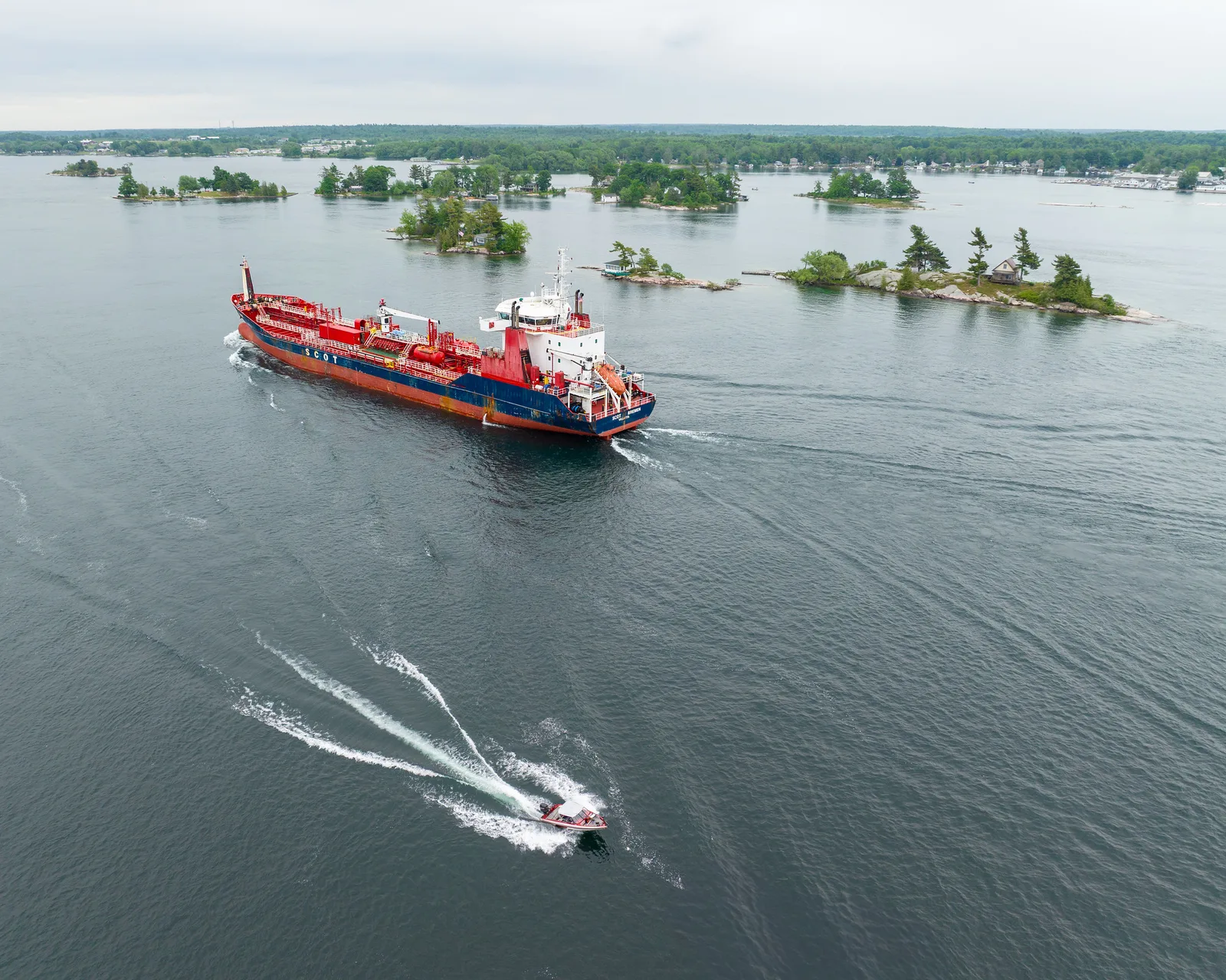 Aerial view of a red tanker ship and a small speedboat with a curving white wake crossing a wide river dotted with wooded islands