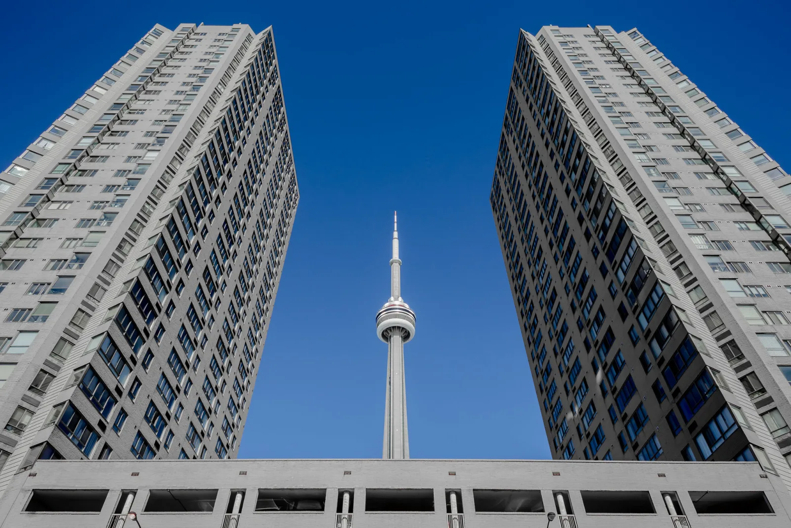 Looking straight up between two tall residential towers that lean inward toward a slender communication tower in the centre, against a blue sky