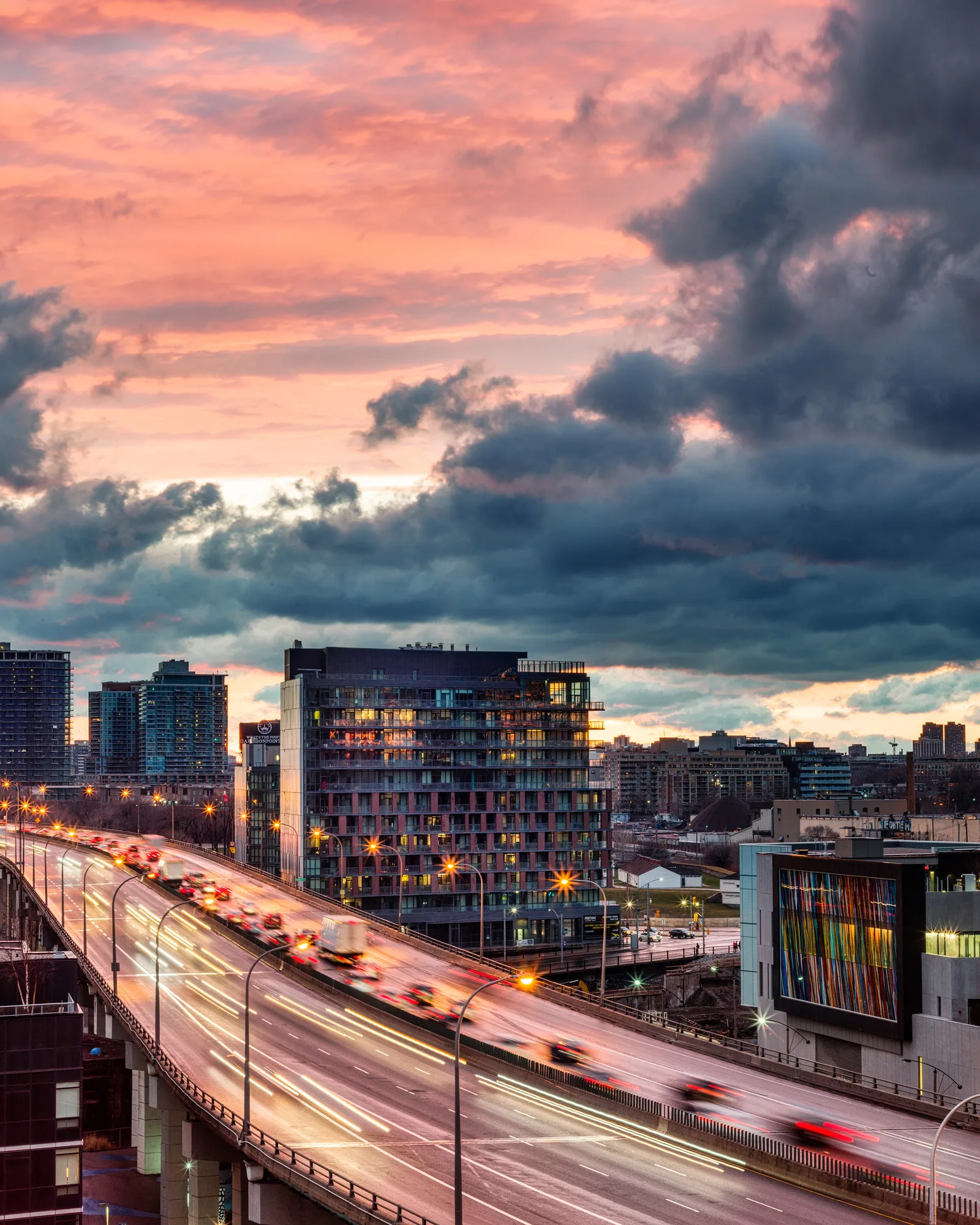 An elevated highway curving through a city at dusk under pink and grey clouds, with cars showing as blurred shapes with short red and white streaks