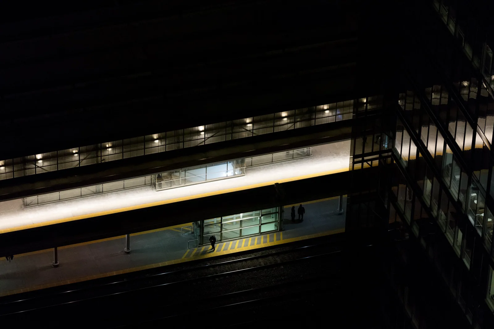 A railway station platform seen from high above at night, lit by rows of artificial ceiling lights, with a few small figures standing near the tracks