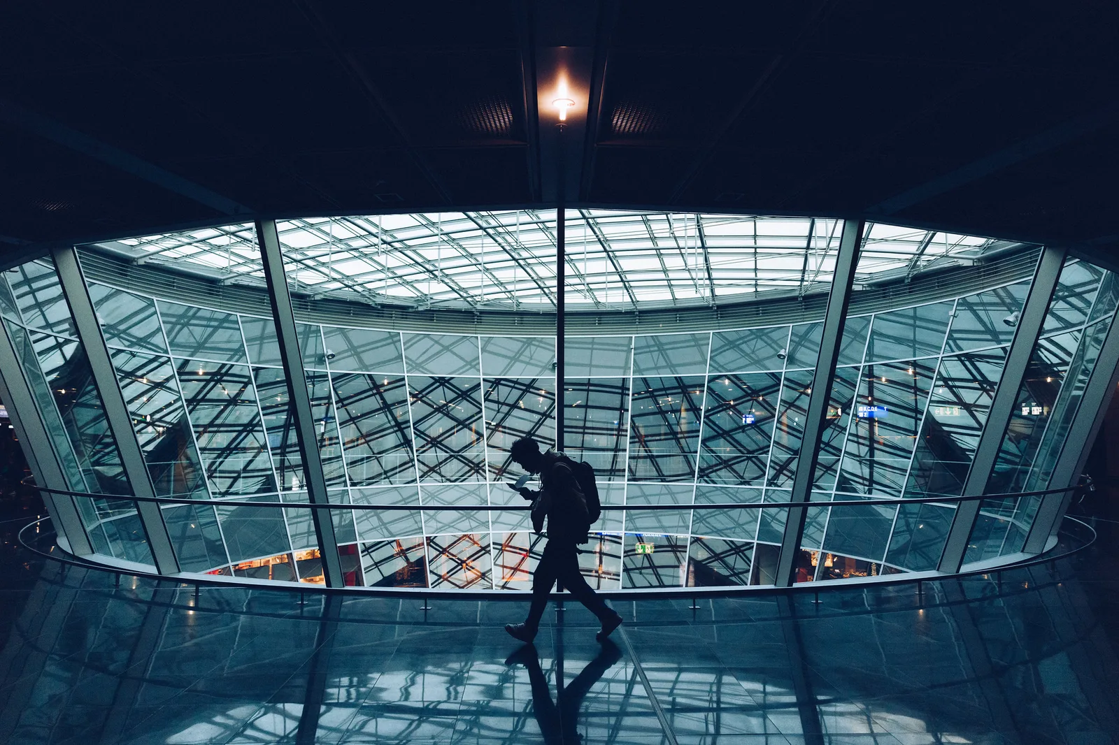 The silhouette of a traveller with a backpack walking past a large curved glass atrium inside an airport terminal at night