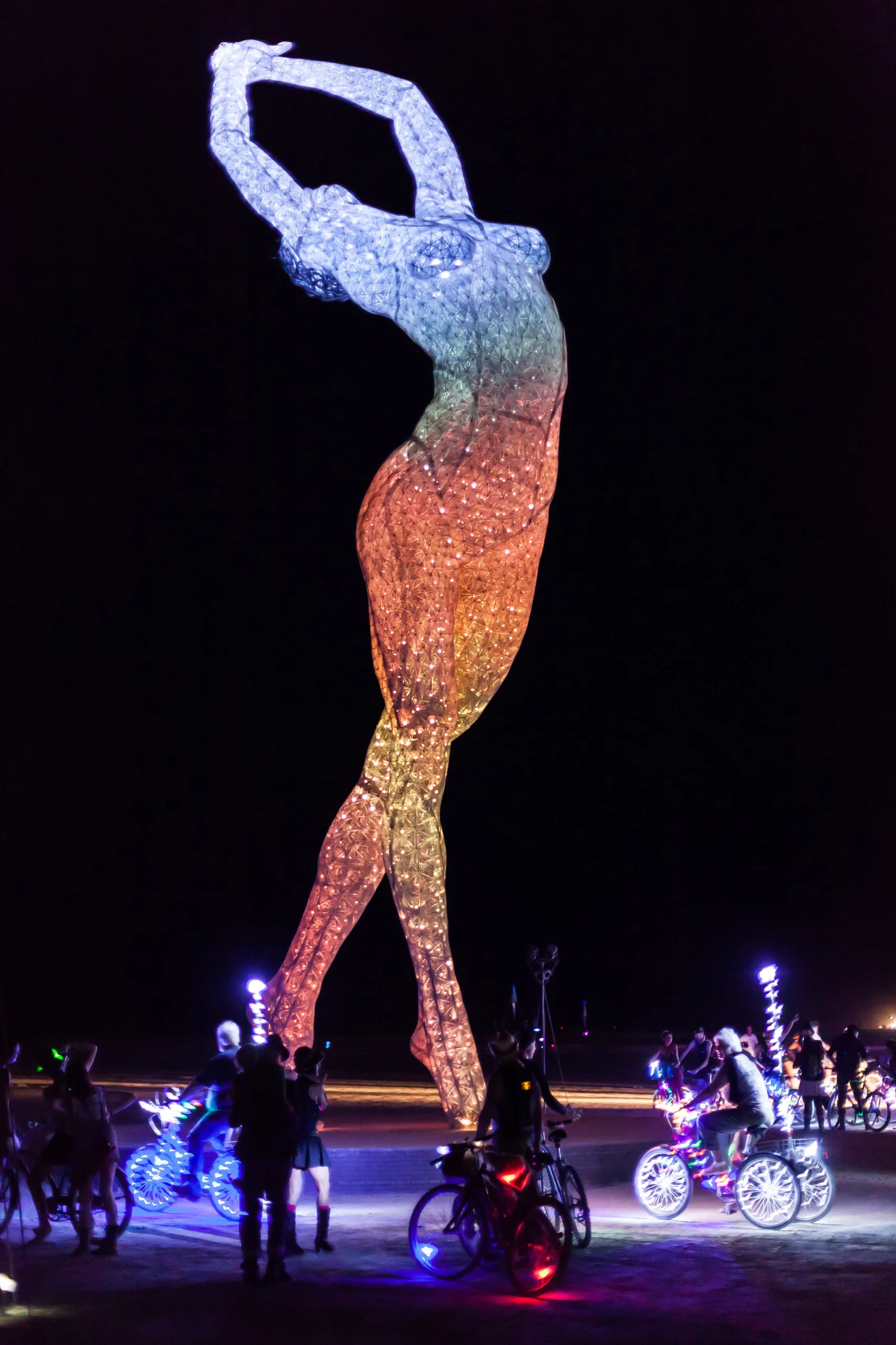 A glowing wireframe sculpture of a dancing figure at night, lit by a nearby light source while the surrounding desert falls into darkness