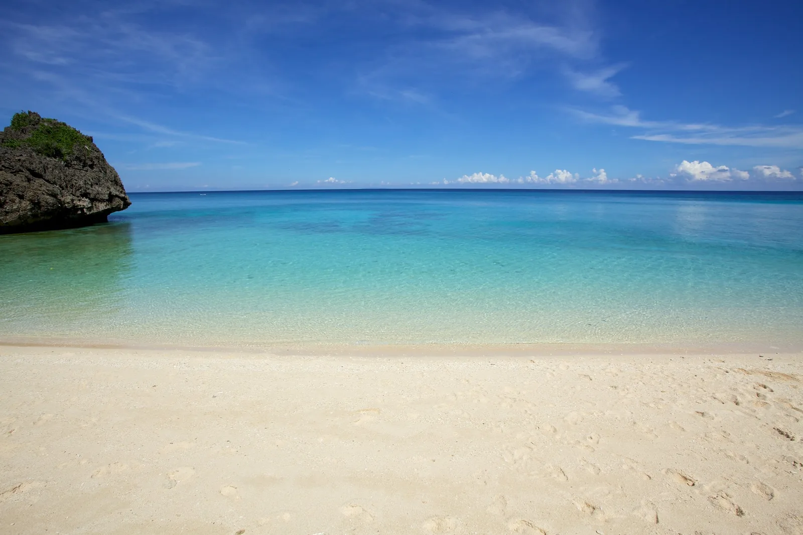 Clear turquoise shallow water over pale sand under a deep blue sky, with white sand in the foreground and a dark rock outcrop at the left
