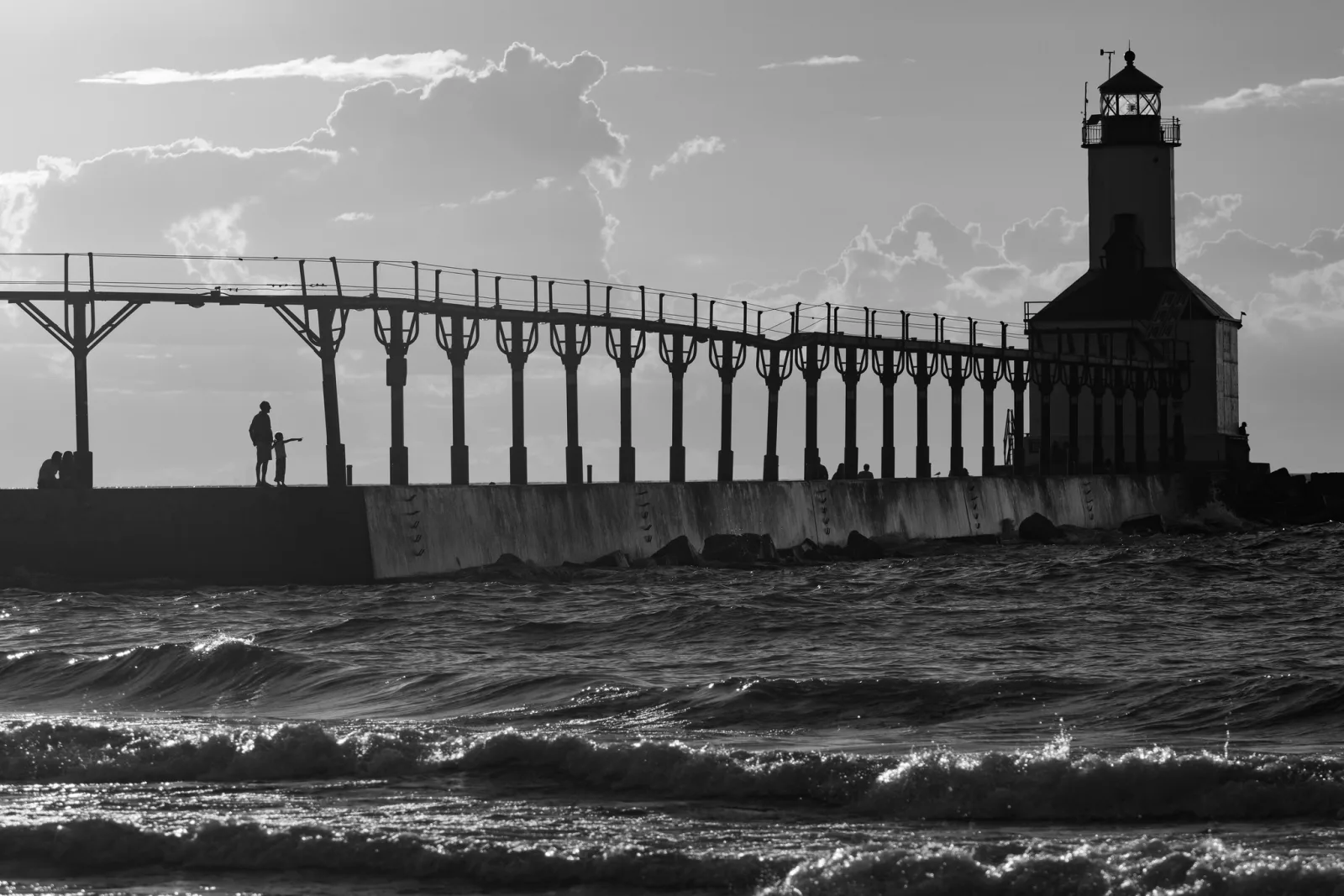Black and white photo of a lighthouse at the end of a long pier with an elevated catwalk, two small figures standing on the pier and waves in the foreground