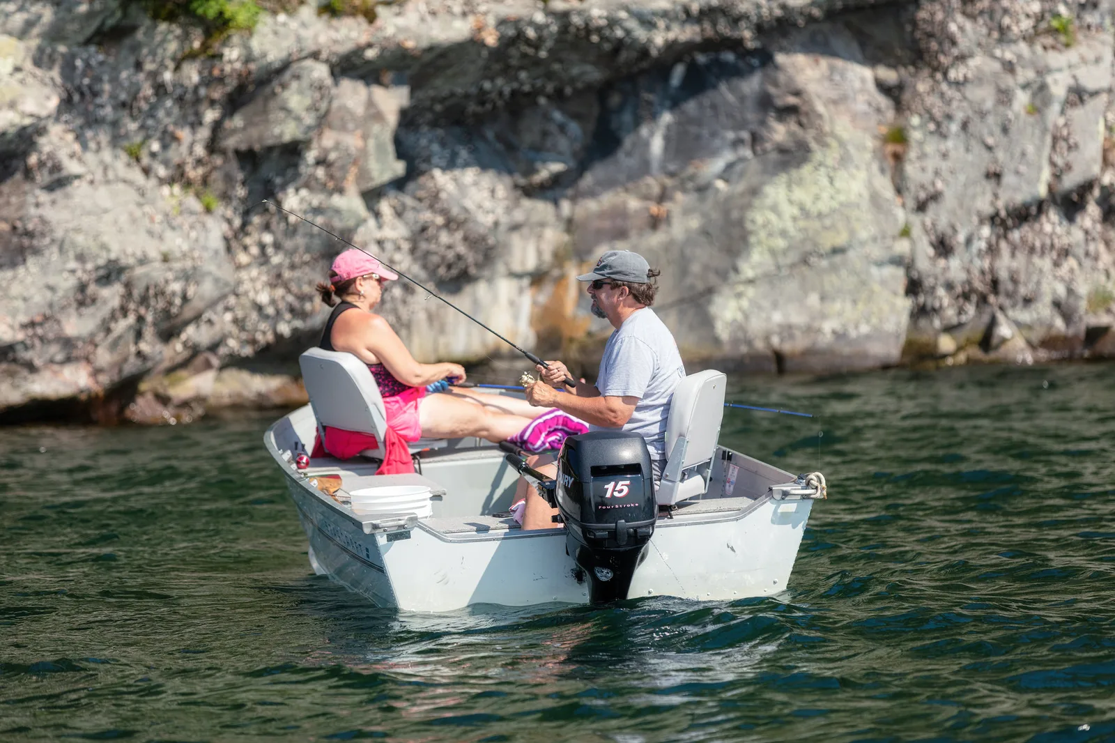 Two people in sun hats fishing from a small white motorboat on green water in bright sunshine, with a rocky shoreline behind them